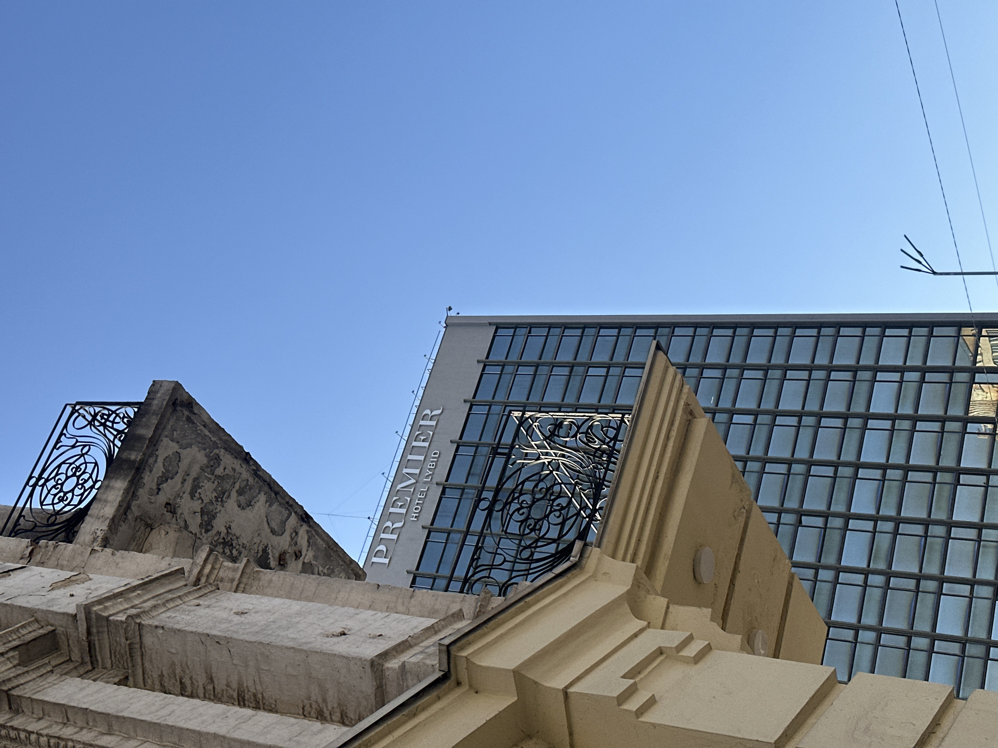The image shows an upward view of architecture in Kyiv, Ukraine. A modern building with a reflective glass facade, identified as "Premier Hotel Lybid" by the signage, is partially visible. Overlapping the modern building are sections of older, possibly historic, stone structures with ornate details. The stone structures feature decorative ironwork and weathered surfaces. The sky is clear and blue. An antenna and wires are visible near the top right corner.