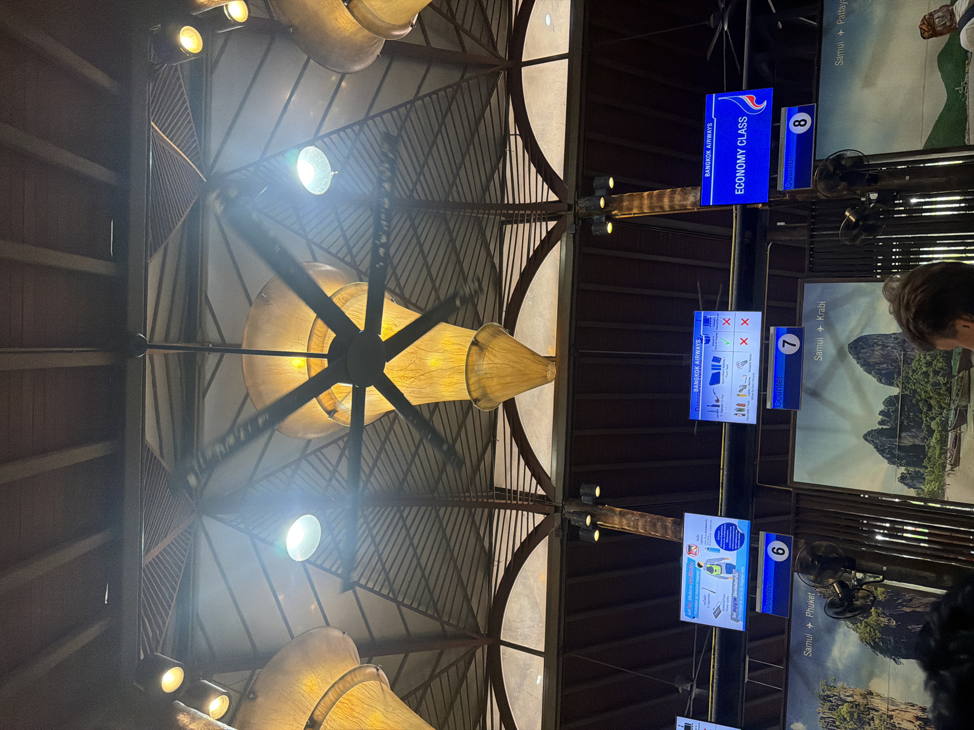 The image shows an indoor scene, likely an airport check-in area in Thailand, given the location context. The perspective is looking upwards towards the ceiling, revealing a distinctive architectural design with large, conical lamps casting a warm glow. The ceiling structure is made of metal beams and what appears to be a fabric or translucent material, creating a patterned effect.
Dominating the scene are several digital information screens and signs mounted on a dark, industrial-looking structure that supports the lamps. One prominent screen displays "BANGKOK AIRWAYS ECONOMY CLASS" in bright blue and white lettering, along with the number "8" indicating a counter. Another screen shows "Samui Phuket" and "Samui Krabi," suggesting flight destinations. A screen closer to the bottom displays "BANGKOK AIRWAYS" with icons indicating prohibited items like "E-cigarettes" and "Spare Lithium Batteries," and permitted items like a power bank. Another screen, with the number "6," shows a graphic of a person with items listed below, possibly related to baggage rules. The text "Samui Phuket" is also visible on a sign.
The overall impression is of an organized, modern airport environment. The lighting suggests it's likely daytime or well-lit interior, as there's no indication of natural light contributing significantly to the illumination. No people are clearly identifiable as subjects in the image, although the presence of the screens and signage implies a functional check-in area. The focus is primarily on the airport infrastructure and information displays.