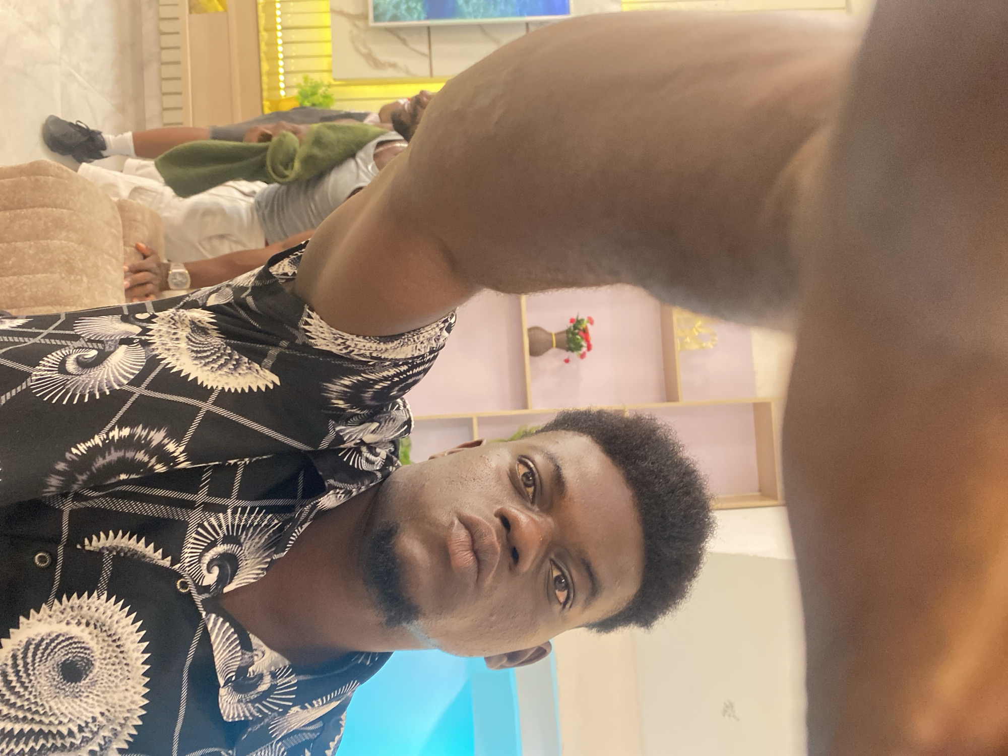 A close-up, angled indoor shot features a young man with short, dark curly hair, a slight beard, and wearing a patterned black and white shirt. His face is the focal point, displaying a neutral expression. The man appears to be taking a selfie. In the background, another person lies on a couch, partially obscured by a green blanket, and another person is visible near the wall. Behind the main subject, there's a wall shelf with a vase of red flowers and a blue light fixture. The location is Unknown, Unknown.