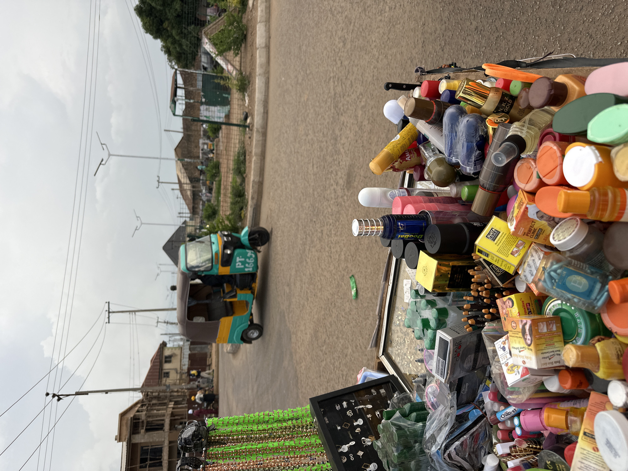 In Bukuru, Nigeria, a roadside stall displays a variety of beauty products and accessories. The stall is filled with numerous bottles, creams, and containers, including hair color boxes, lotions, and various cosmetics. In the foreground, the edge of a stall is visible, laden with these items. Behind the stall, a street scene unfolds with a teal and yellow auto-rickshaw in the middle ground, its license plate reading "PT 16634." Further in the background, construction is underway on a building, and other vehicles are present on the street. The sky is overcast, suggesting a cloudy day.