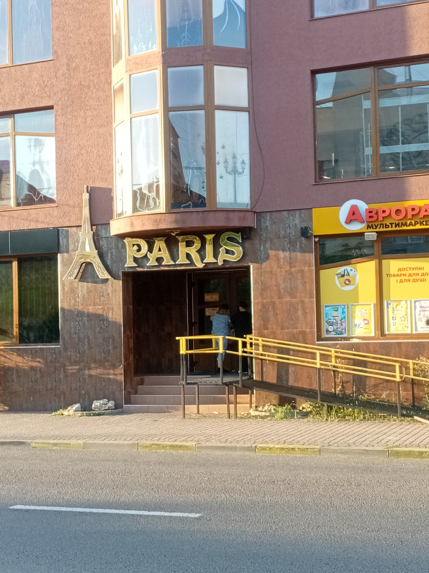 The image shows the facade of a building in Tiachiv, Ukraine, on a sunny day. The building has a brown textured exterior with large windows. On the left side of the entrance, there is a golden Eiffel Tower replica attached to the wall. Above the entrance, the word "PARIS" is displayed in large, golden letters. To the right of the "PARIS" entrance, there is a storefront with a yellow facade and the word "АВРОРА" in red letters, with "МУЛЬТИМАРКЕТ" written below it in smaller text. Below the "АВРОРА" sign, there are several posters and displays visible through the window. A ramp with yellow railings leads up to the entrance, and a set of stairs is also visible. Two people are standing near the entrance to "PARIS". The street in front of the building is paved, and a sidewalk with a yellow curb separates it from the building. Some vegetation is growing near the base of the building.