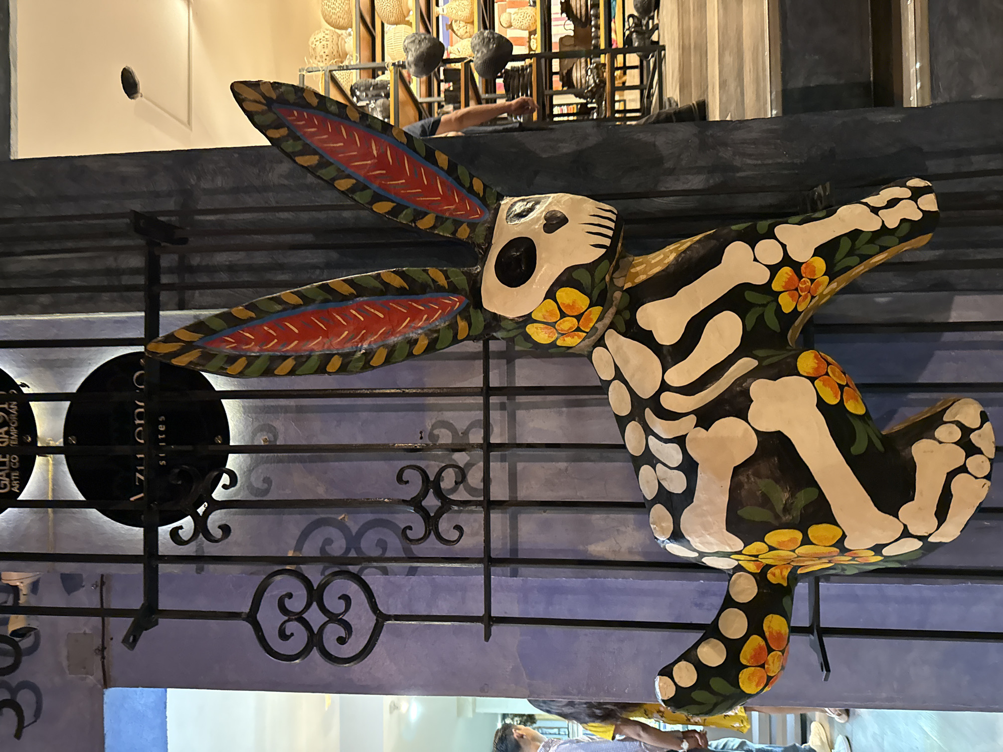 A horizontal, close-up shot shows a painted wooden rabbit skeleton hanging on an ornamental metal fence in Oaxaca City, Mexico. The rabbit is black with white bone shapes and orange and yellow flowers. The ears are decorated with red, blue, and yellow lines. The fence is dark blue, and the wall behind it is light purple. A person’s arm can be seen reaching into the frame from above, and other objects such as woven baskets are visible on a shelf in the background. There is a sign with illegible writing hanging on the fence to the left of the rabbit. Part of a person is visible at the very bottom of the frame.
