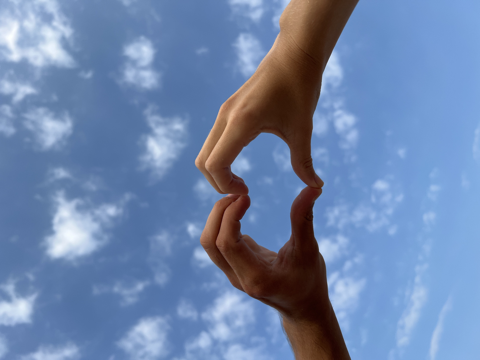 Two hands, one above the other, are positioned against a blue sky with scattered white clouds, forming a heart shape with the fingers. The lower hand is more tan, while the upper hand has a paler skin tone. The location is not identifiable as Nam Natural Heritage Area Trang, Vietnam based solely on the visual elements of the image.