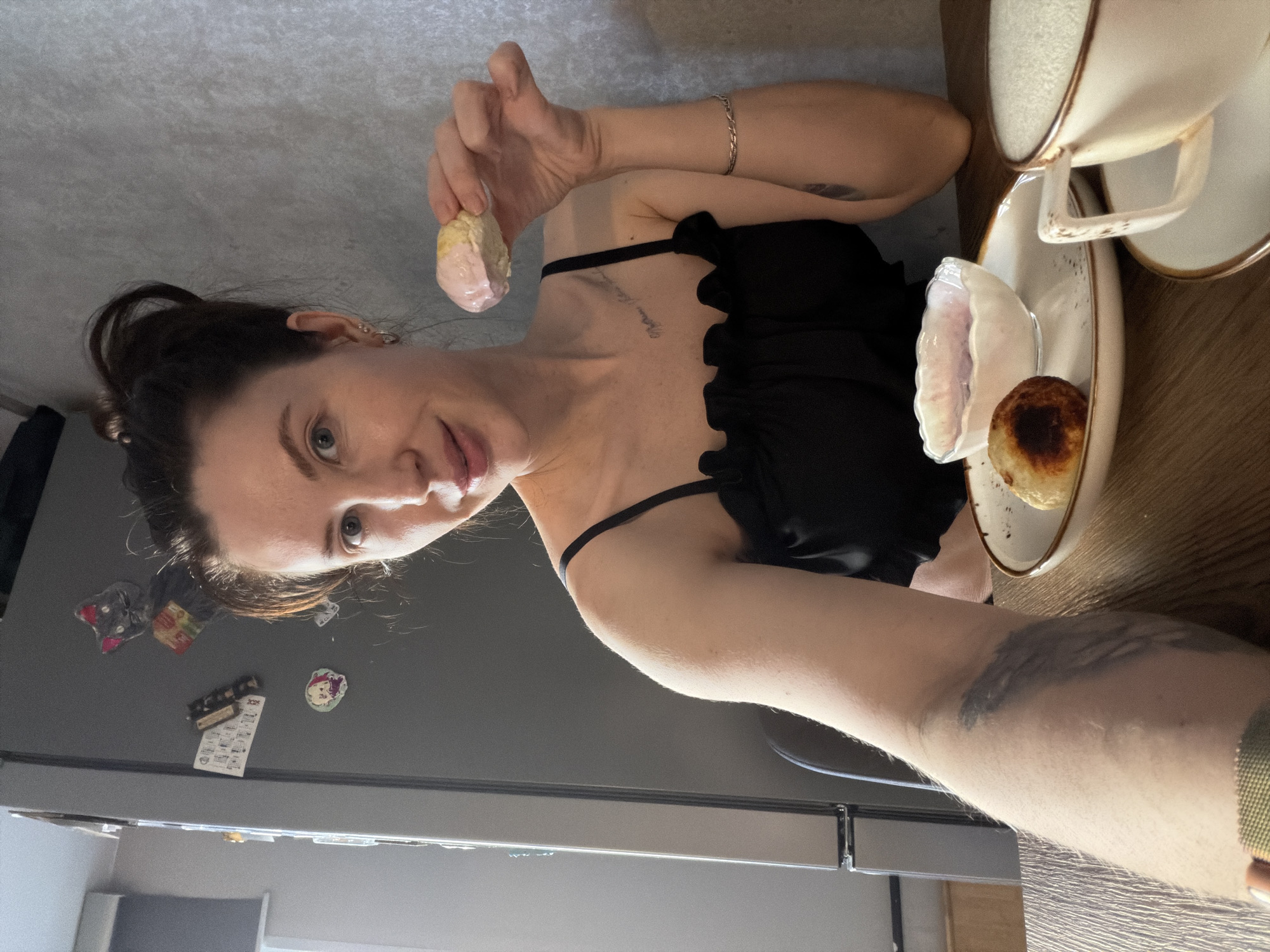 A woman is taking a selfie in an indoor setting. She holds a pastry in her right hand. There is a plate with two other pastries. The woman wears a black top and has a tattoo on her arm. The background includes a refrigerator adorned with magnets. The setting appears to be a kitchen in a residence. There is no information to identify the location as Perm, Russia.