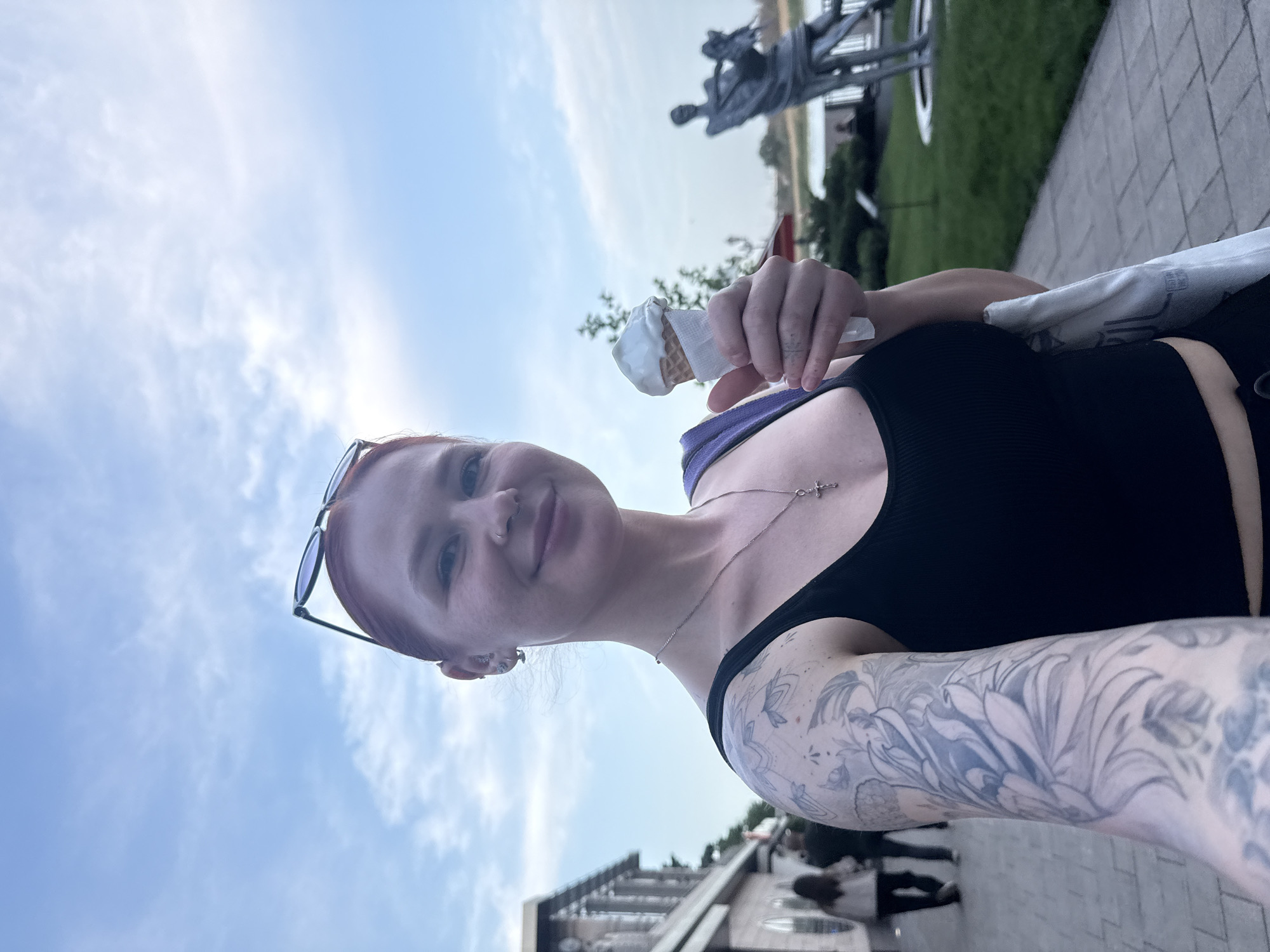 A young woman is holding an ice cream cone in front of her while outdoors. She is wearing a black tank top, sunglasses on her head, and a silver cross necklace. She has tattoos on her left arm. In the background, there are some sculptures and green foliage. Based on the location of these sculptures, the image appears to be taken in Rostov-on-Don, Russia. The sky is visible in the background, with light clouds.