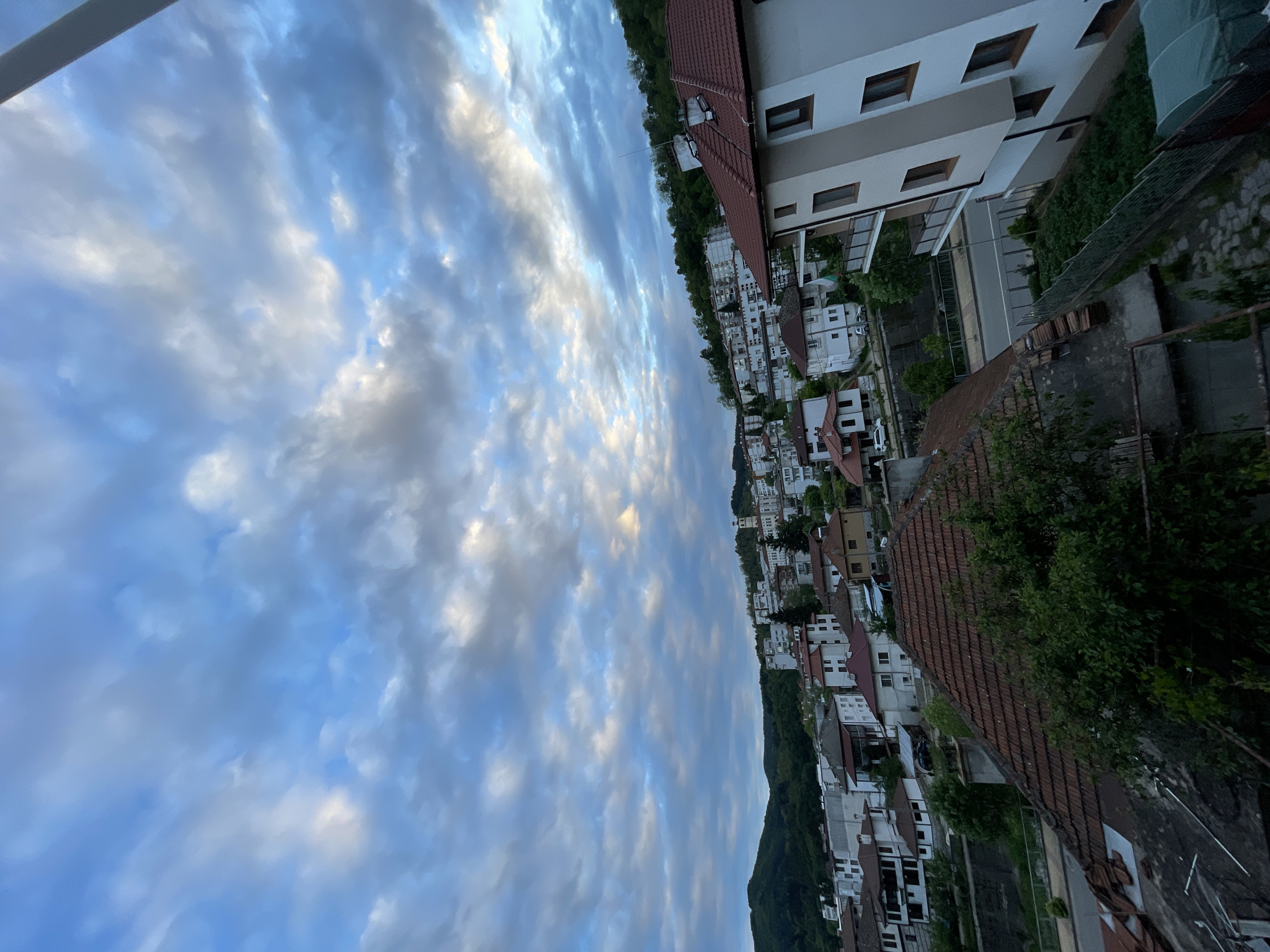 The image shows an aerial view of Zlatograd, Bulgaria. The foreground features a cluster of buildings with red tile roofs. White buildings are seen along the base of a tree-covered hillside. Above the buildings, a cloudy blue sky fills much of the frame.
