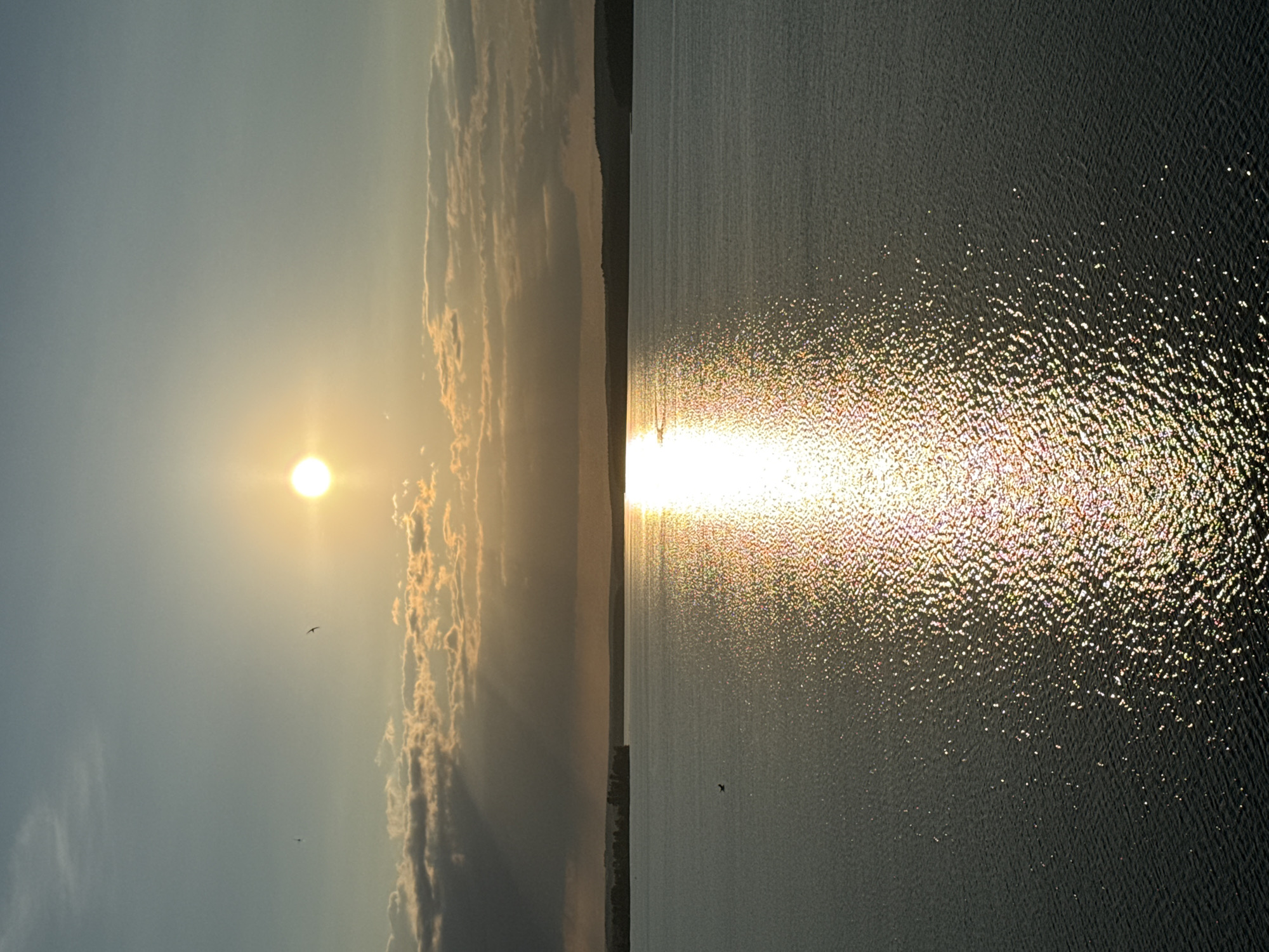 This image depicts a serene sunset over a wide body of water, likely the Votkinsk Reservoir, near Votkinsk, Russia. The sun, a bright, almost white orb, hangs low in a pale blue sky, casting a brilliant, shimmering reflection across the water's surface. Streaks of light emanate from the sun, piercing through scattered, wispy clouds that are tinged with the warm hues of dusk. The horizon is marked by a dark, silhouetted line of trees and hills. The water itself is calm, with gentle ripples that catch the light, creating a sparkling effect. In the distance, a small, dark landmass, possibly an island or peninsula, juts out into the reservoir. The overall atmosphere is one of peaceful tranquility and natural beauty, characteristic of a sunset in this region.