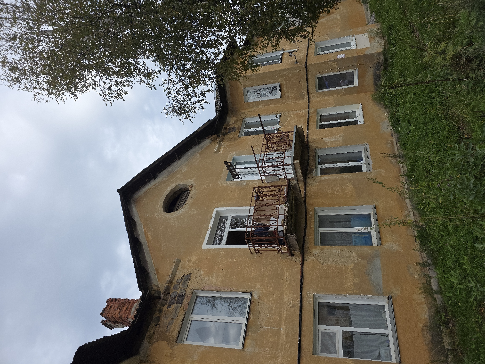 The image shows a two-story building with a weathered, mustard-yellow facade. The building appears to be in disrepair, with peeling paint and visible brickwork in some areas. There are multiple windows with white frames, some of which have curtains. A small, rusted metal balcony juts out from the second story, with clothing hanging on its railing. A brick chimney is visible on the roof to the left, and a large tree with green foliage partially obscures the right side of the building. The ground in front of the building is covered in overgrown green grass and some weeds. The sky is overcast with a light gray cloud cover. The scene is likely situated in a residential area of Magnitka, Russia, given the architectural style and general appearance. There are no people visible in the image.