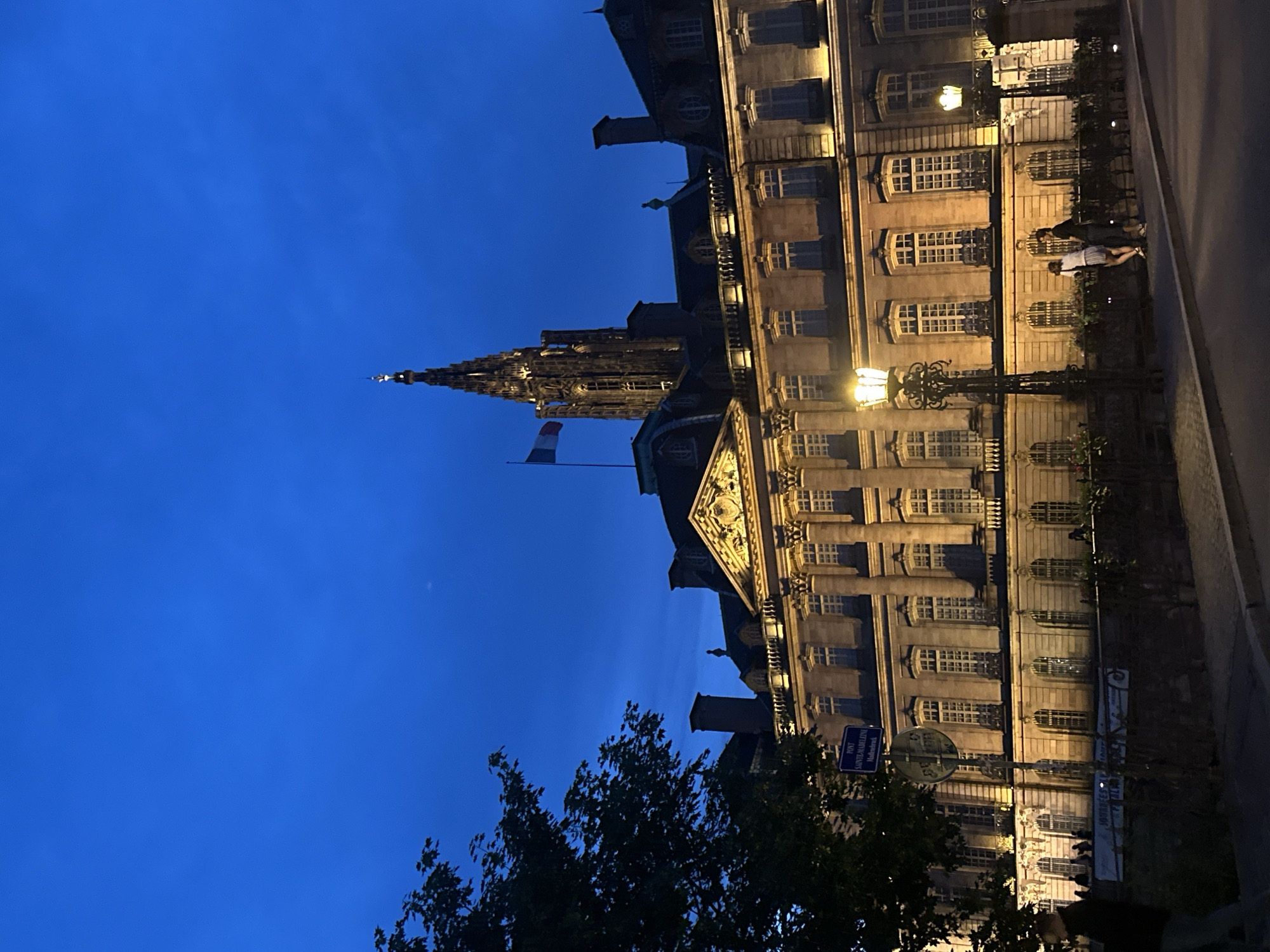 The image depicts an urban scene at dusk or night, characterized by a deep blue sky and illuminated historic architecture. In the foreground, silhouetted trees frame the left side of the composition.
Dominating the left-central portion of the image is the majestic, intricate spire of a Gothic cathedral, piercing the dark blue sky. Below and to the left of the spire, a French flag (blue, white, red) flies from a flagpole.
To the right, a grand, multi-story building with numerous windows extends horizontally across the frame. This building is brightly illuminated by artificial lights, highlighting its classical architectural details, including ornate carvings above the main entrance and along its facade. Several street lamps with decorative ironwork are positioned in front of this building, casting additional light onto the scene.
On the pavement in front of the illuminated building, a few people are visible, appearing to walk or stand. One person, wearing a light-colored top and dark pants/skirt, is clearly discernible.
Notable details include the clear, dark blue sky, indicating either twilight or nighttime, with a faint crescent moon visible in the upper right portion of the sky. The illumination of the buildings suggests they are public structures or significant landmarks.
A blue street sign is visible in the lower-middle left, displaying text in both French and German: "PONT SAINTE-MADELEINE" and "Madeleinebrück". This indicates the location is the Pont Sainte-Madeleine (St. Madeleine Bridge). Given the architectural style, the French flag, and the bilingual street sign, the scene is definitively set in Strasbourg, France, with the prominent spire likely belonging to the Strasbourg Cathedral.