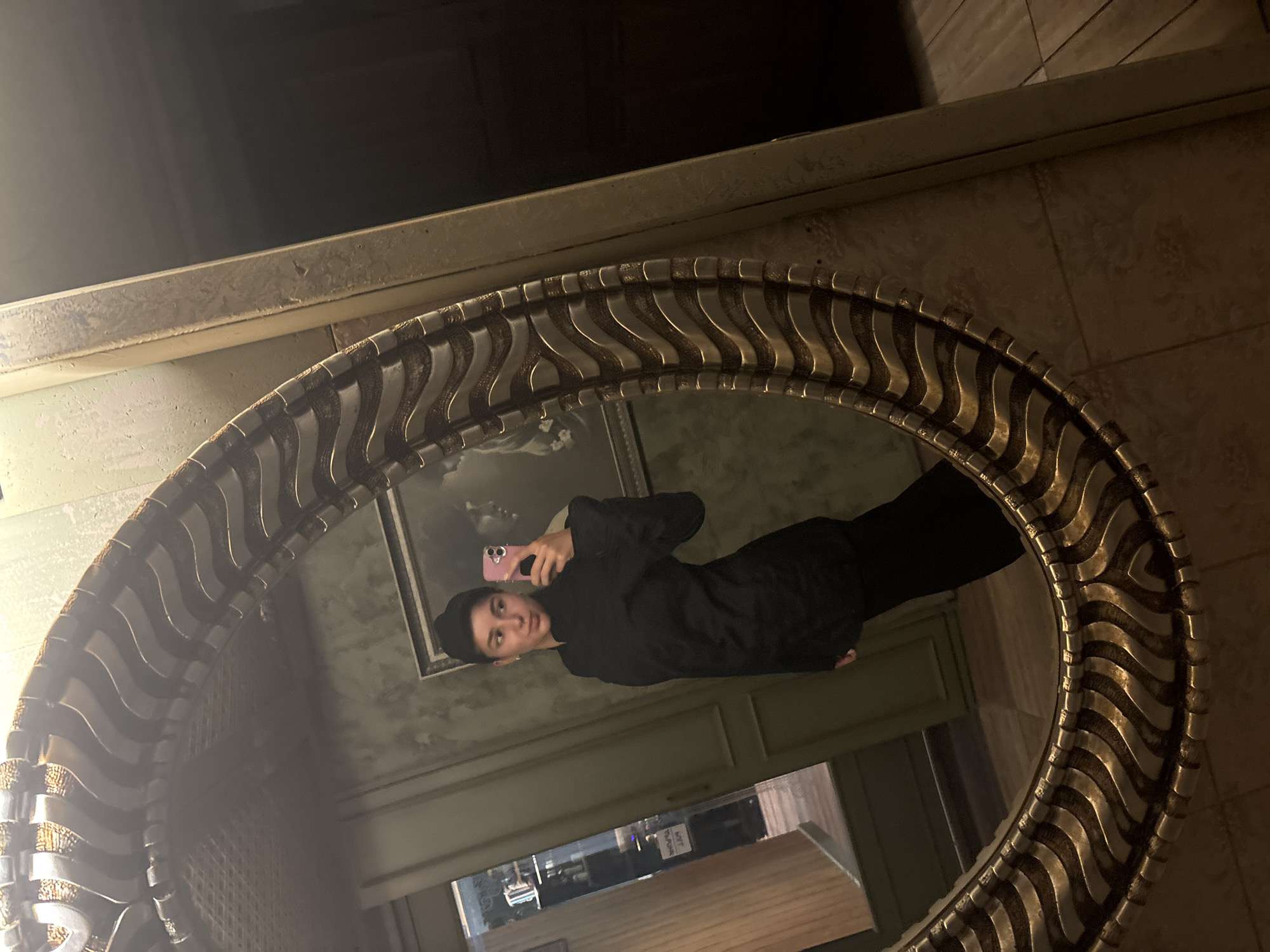 A person takes a mirror selfie in a room. The mirror is oval with an ornate, gold-colored frame. The person is wearing black clothes and holding a smartphone. The background reflects a room with a painting, window, and furniture. Outside the window, city buildings are visible. A white sign with what seems to be black letters is seen in the reflection. The location appears to be Veliko Tarnovo, Bulgaria based on building style.