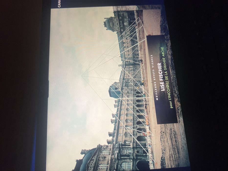 The image displays a photograph of a screen exhibiting an outdoor scene. The depicted location is the Cour Napoléon of the Louvre Museum in Paris, France.
The central subject is a large, temporary art installation composed of numerous white lines forming a transparent, wireframe pyramid. This structure creates an optical illusion, seemingly rising from a disturbed ground area in the foreground and converging to an apex against the backdrop of the historic palace.
The environment consists of the grand, classical Louvre Palace, featuring multi-story facades with numerous windows, intricate architectural details, arched ground-floor passages, and characteristic Mansard roofs. The palace structures extend across the entire background. The ground immediately surrounding the installation appears to be a construction or renovation site, consisting of bare, brown earth. A few orange cones or barriers are visible within this area, indicating ongoing work.
Several small, indistinct human figures are present in the mid-ground near the palace, but no specific actions or interactions are clearly discernible.
A dark, rectangular graphical overlay, likely part of a broadcast presentation, occupies the lower right foreground. It displays white text: "MEILLEURS EFFETS VISUELS" (Best Visual Effects), "LISE FISCHER," and "POUR L'INCONNU DE LA GRANDE ARCHE" (For the Unknown of the Grande Arche). A partial "CANA" is visible in the upper right corner, likely from a channel logo.
The sky is overcast and light-colored, providing soft, diffuse illumination to the scene. The image itself shows signs of being a photograph of a screen, including a darker frame around the edges and slight pixelation.