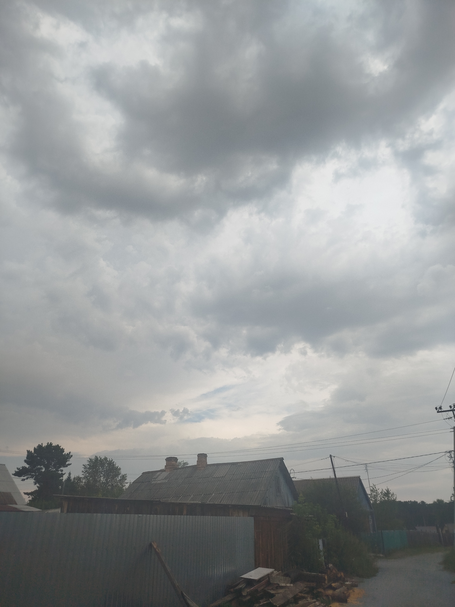The image captures a cloudy sky over a rural residential area in Zavodoukovsk, Russia. In the foreground, a corrugated metal fence partially obscures a view of several wooden structures, including a house with a grey, corrugated roof and two chimneys. To the left, a portion of another house with a grey roof is visible. Behind these structures, trees and vegetation are present. Overhead, power lines are strung between utility poles, indicating the presence of infrastructure. The overall atmosphere is overcast, suggesting a gray or possibly rainy day. The scene appears to be a quiet, undeveloped area.