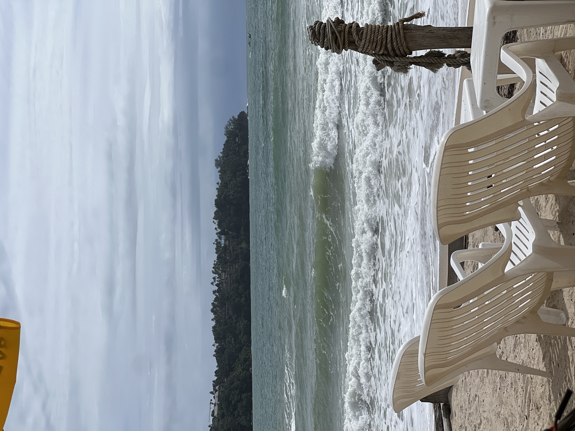 The image depicts a sandy beach scene under an overcast sky. The upper third of the frame shows a light grey, cloudy sky with horizontal cloud formations. The middle ground features a vast expanse of greenish-teal ocean, characterized by numerous breaking waves cresting with white foam, especially prominent closer to the foreground. On the left side of the distant horizon, a dark green, heavily forested headland is visible, dotted with buildings featuring light-colored roofs, some appearing to be residences or resort structures. A construction crane is discernible above the tree line on the far left of the headland. A small, dark, elongated boat is present on the water towards the far right in the distance.
The immediate foreground consists of light brown sand. Two white plastic slatted chairs are positioned on the sand, facing the ocean, with a third chair or table partially visible on the far right. A small, square white plastic table sits between the two chairs on the right. To the right of these furnishings, closer to the water, a weathered wooden post is embedded in the sand. This post is heavily wrapped with thick, tan-colored rope, coiled densely around its upper section, with several loose rope ends hanging downwards. The incoming tide creates a line of white foam along the shoreline, reaching the base of the chairs and the wooden post. A partial view of a yellow object, possibly an umbrella or canopy, is present in the top left corner of the frame. No individuals are visible in the scene.