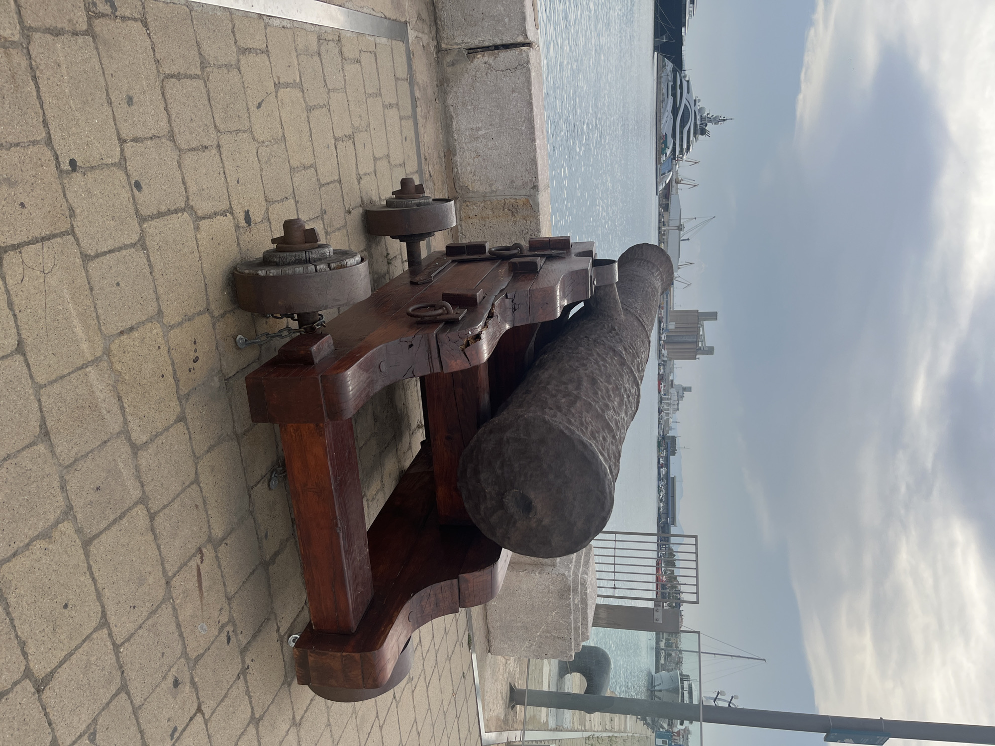 The image shows a historical cannon on a stone-paved walkway next to a body of water. The cannon is made of wood and metal, with a long, cylindrical barrel. It is mounted on a wooden carriage with wheels. In the background, there are several boats and ships, as well as buildings and cranes. The sky is cloudy. The scene is consistent with a maritime location, possibly a harbor or port. This cannon is located in the port of Tarragona, Spain, near the Roman Amphitheatre and the Mediterranean Sea.