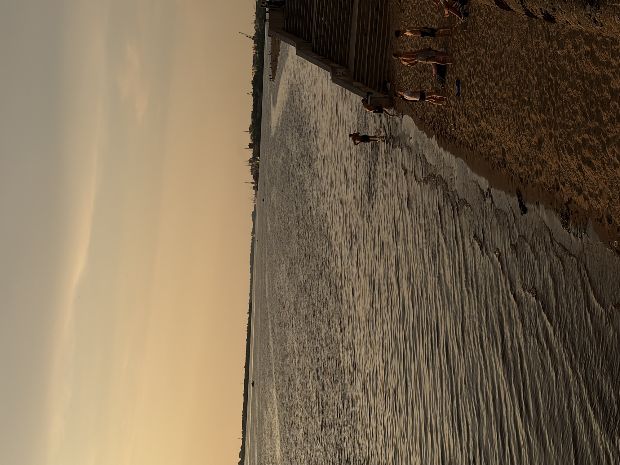 The image depicts a wide, calm river with gentle waves lapping against a sandy beach. In the background, the silhouette of the city of Arkhangelsk, Russia, is visible with industrial cranes and buildings. Several people are on the beach, some wading into the water, while others stand or sit on the sand. A concrete staircase leads down to the beach. The sky is a soft, warm hue, suggesting either sunrise or sunset.