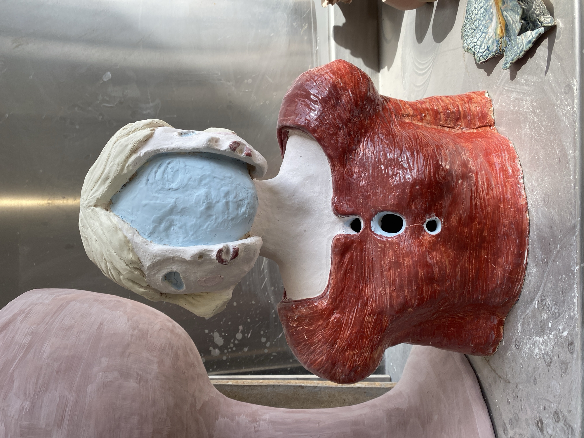 The image shows a partial, apparently unfinished ceramic sculpture. The sculpture consists of a red, roughly head-shaped form with holes for eyes, attached to a white neck leading to an open mouth with light blue inside. The mouth and the neck are a contrasting, lighter color than the red head. The sculpture is resting on another, larger, pink ceramic form, which appears to be part of the same sculpture but is less detailed. The background is a stainless-steel surface with water stains. Other ceramic pieces can be seen partially in the background. There is no evidence to suggest a specific location within Saint-Ouen-sur-Seine or any other city.
