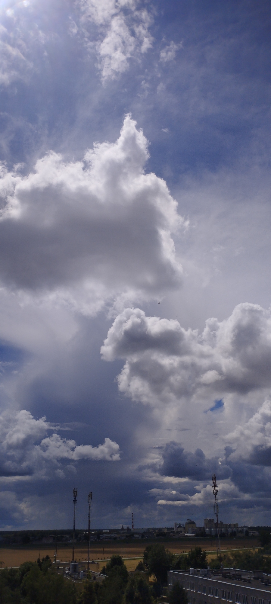 A wide shot captures a dramatic sky filled with a mix of light and dark cumulonimbus clouds. The sun is visible in the upper left corner, casting a bright light on the scene. In the middle ground, a flat landscape stretches across the horizon, featuring fields of dry grass and scattered trees. Several prominent structures rise from the landscape, including communication towers and industrial buildings. Two tall, dark communication towers stand side-by-side in the left portion of the frame, while another similar tower is visible on the right. In the distance, industrial facilities with smokestacks and large silos are discernible. The bottom of the frame shows the roof of a building and the tops of trees. The overall impression is one of a vast, open sky over an industrial area, with the weather creating a sense of depth and intensity. The location is Skidzieĺ, Belarus.
