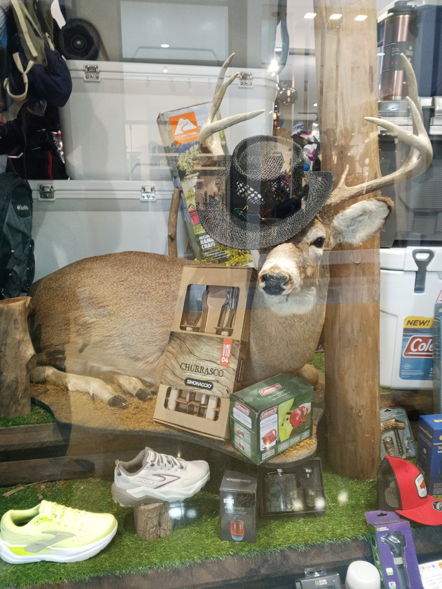The image displays a retail store window filled with various outdoor and sporting goods. In the foreground, two pairs of athletic shoes, one neon yellow and the other a light gray, are showcased on a faux grass surface. A taxidermied deer head, adorned with a black cowboy hat, is positioned prominently behind the shoes. Various boxes and packages containing camping and hunting accessories, including coolers, a quick pump, and a hatchet, are arranged around the deer. A large, white cooler is visible in the background, along with stacks of what appear to be additional storage containers. The scene is set against a backdrop of what seems to be a store interior, with artificial lighting illuminating the products.