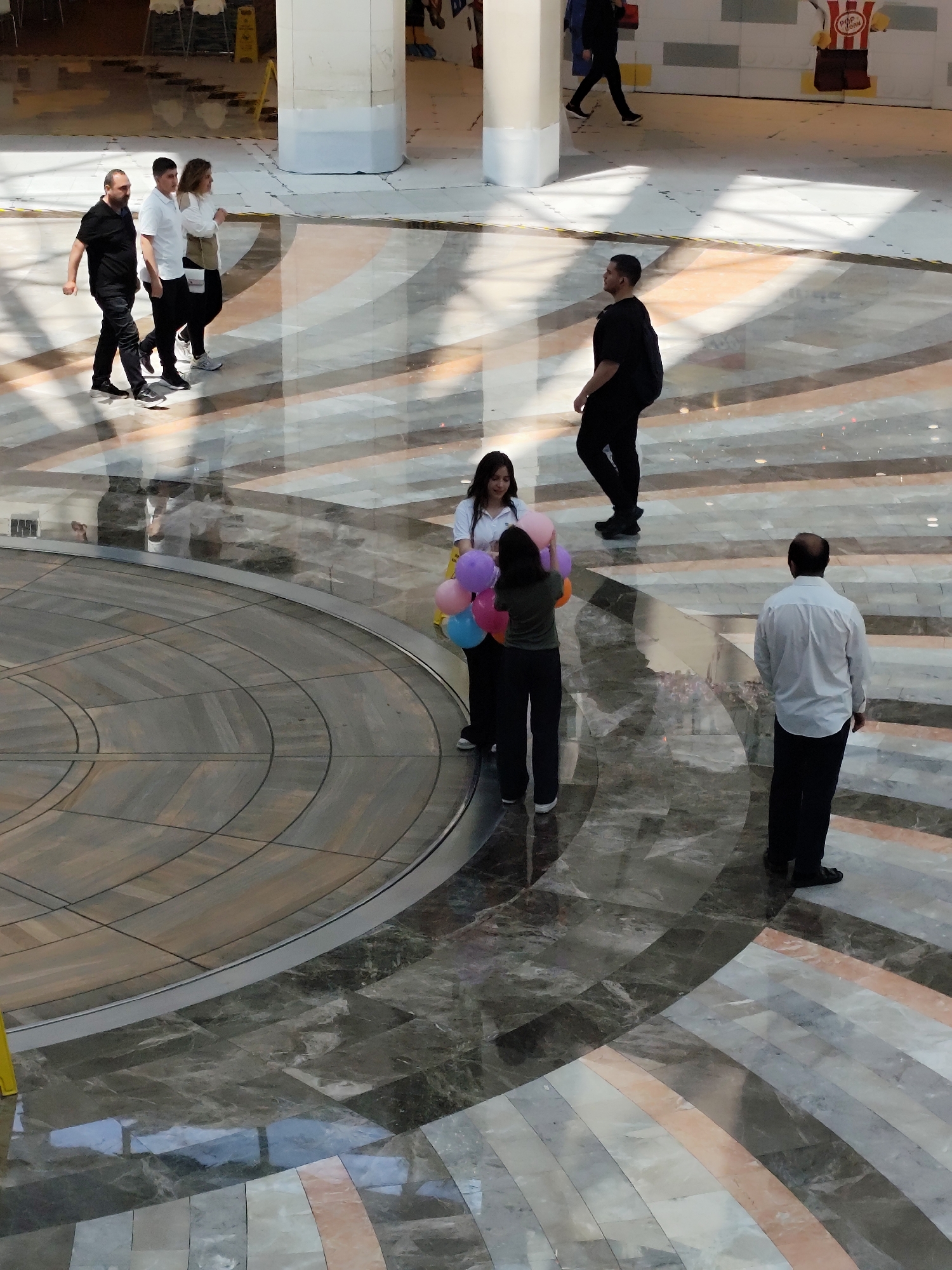 Captured from an elevated angle within a multi-story building in Sarıyer, Turkey, the image displays a scene on a patterned marble floor. The scene is illuminated by natural light through the building's architecture. Multiple people are walking on the patterned marble floor with light reflections. A woman holds balloons and gives them to another woman. A group of three people walk together, and two individuals are walking separately. The backdrop includes building columns, other people, and a concession stand.