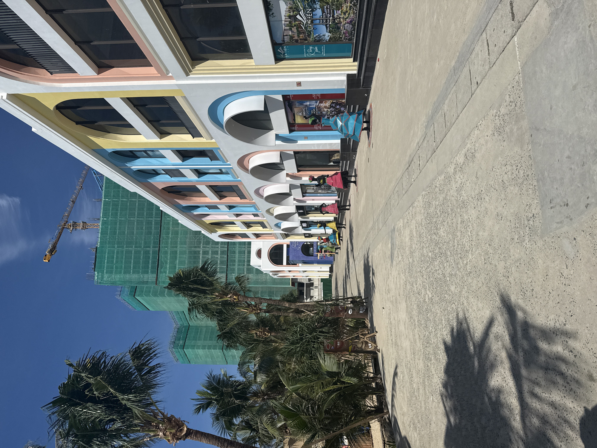 The image depicts a pedestrian walkway in front of a colorful, modern building with arched windows and balconies. The building's facade is painted in pastel shades of yellow, pink, blue, and white. On the left side of the walkway, several palm trees are visible, casting shadows on the ground. In the background, large buildings are under construction, covered in green netting, with a yellow crane extending into the clear blue sky. Several mannequins dressed in colorful, geometric dresses are displayed along the walkway, creating an artistic installation. One of the shops visible has signs in Vietnamese, with the text "LA TIEN VILLA" and a phone number. This scene is consistent with the areas in Nha Trang, Vietnam that are developing new tourist facilities and entertainment venues.