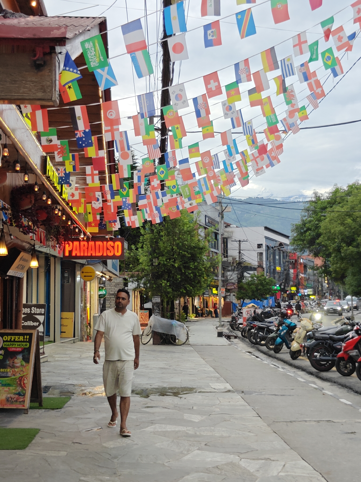 The image captures a street scene in Pokhara, Nepal, likely along Lakeside Road, characterized by a vibrant display of international flags strung across the street, suggesting a tourist-oriented area. A man in a white t-shirt and light-colored shorts is walking on the paved sidewalk from left to right, towards the viewer. He is wearing flip-flops. To his left, a storefront displays a prominent red neon sign that reads "PARADISO." Adjacent to the "PARADISO" sign is an ATM and other shops with goods visible behind glass. A wooden A-frame sign with text and images, including "OFFER!!" and "HAPPY HOUR," is positioned on the sidewalk to the left of the man. The street is lined with parked motorcycles and a few cars. Trees and buildings form the backdrop, with distant mountains visible under an overcast sky, characteristic of the Pokhara valley. The overall atmosphere suggests a lively, possibly commercial or entertainment district in the city.