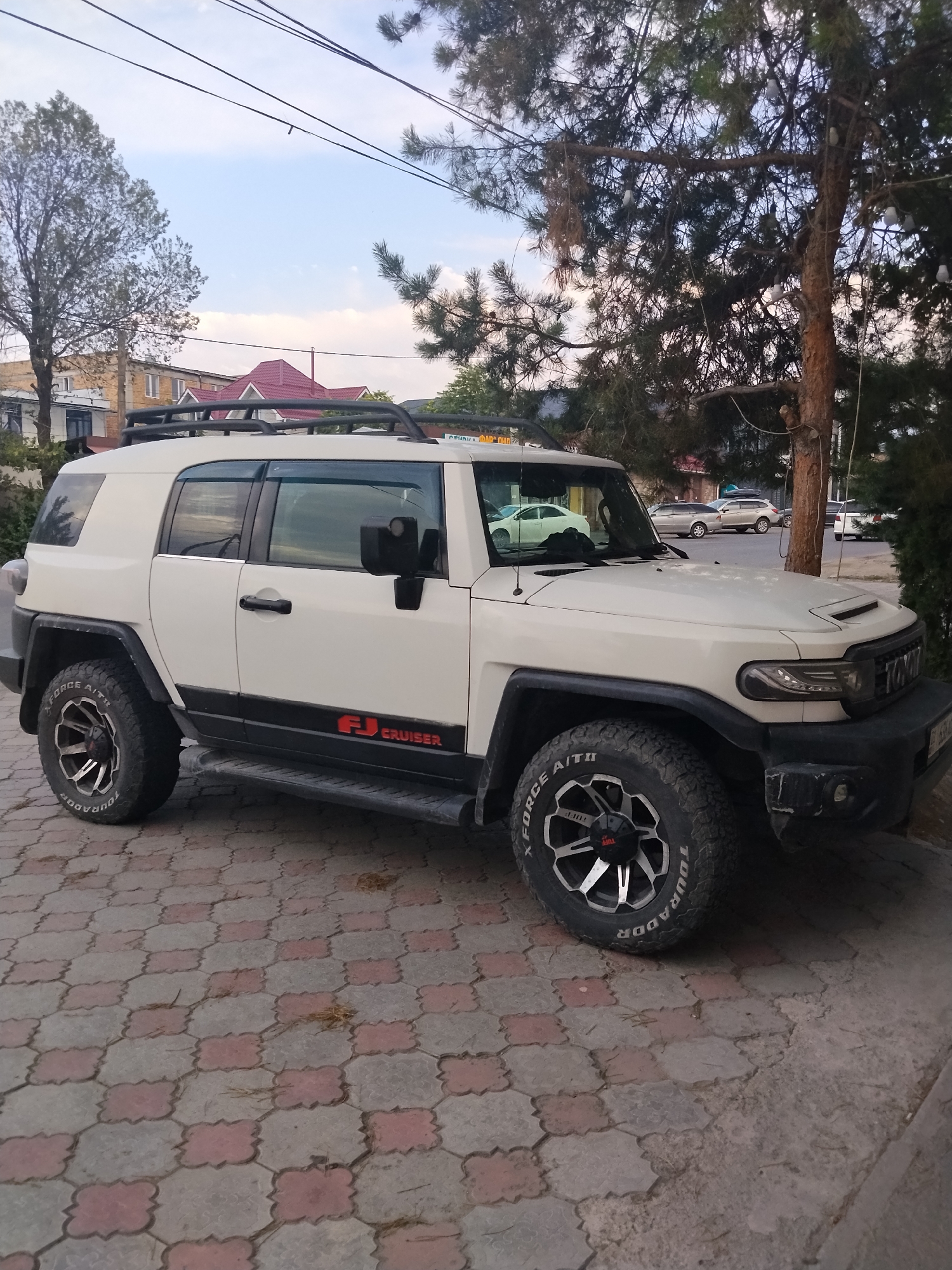 A white 2010-2017 Toyota FJ Cruiser is parked on a cobblestone street, with its left side facing the viewer. The SUV has a roof rack, tinted windows, and distinctive red "FJ CRUISER" badging on its lower body. It is equipped with off-road tires and black rims. The car is parked near trees and buildings, with power lines visible overhead. The sky is cloudy.