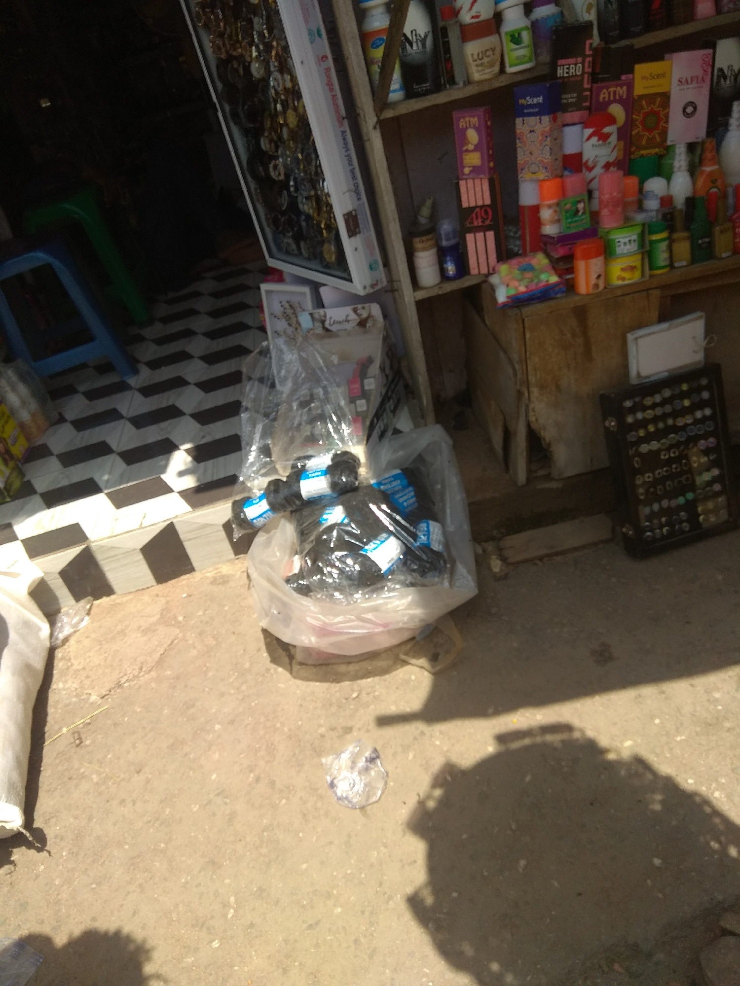 This is an outdoor medium shot of a retail display, likely a street stall, in Jalingo, Nigeria. The primary focus is on shelves stocked with various products. On the top shelf, there are numerous small bottles and containers, some with labels like "NY," "LUCY," and "HERO." Below this, a display features items with prominent "ATM" and "myScent" branding, along with what appear to be gift sets and small boxes. A collection of rings is displayed in a black tray on a lower shelf.
In the foreground, a large clear plastic bag is filled with black items, possibly hair extensions or similar accessories, with blue and white labels visible. A white fabric object is partially visible on the left. The ground is a mix of concrete and rough dirt, with shadows indicating a bright, sunny day. The background shows a patterned tiled floor and part of a stall structure. The overall scene suggests a commercial area where various goods are sold, likely in an open-air market or street-side shop. The bright sunlight and shadows suggest it is daytime, and the abundance of products points to an active retail environment. No people are clearly visible in the frame.