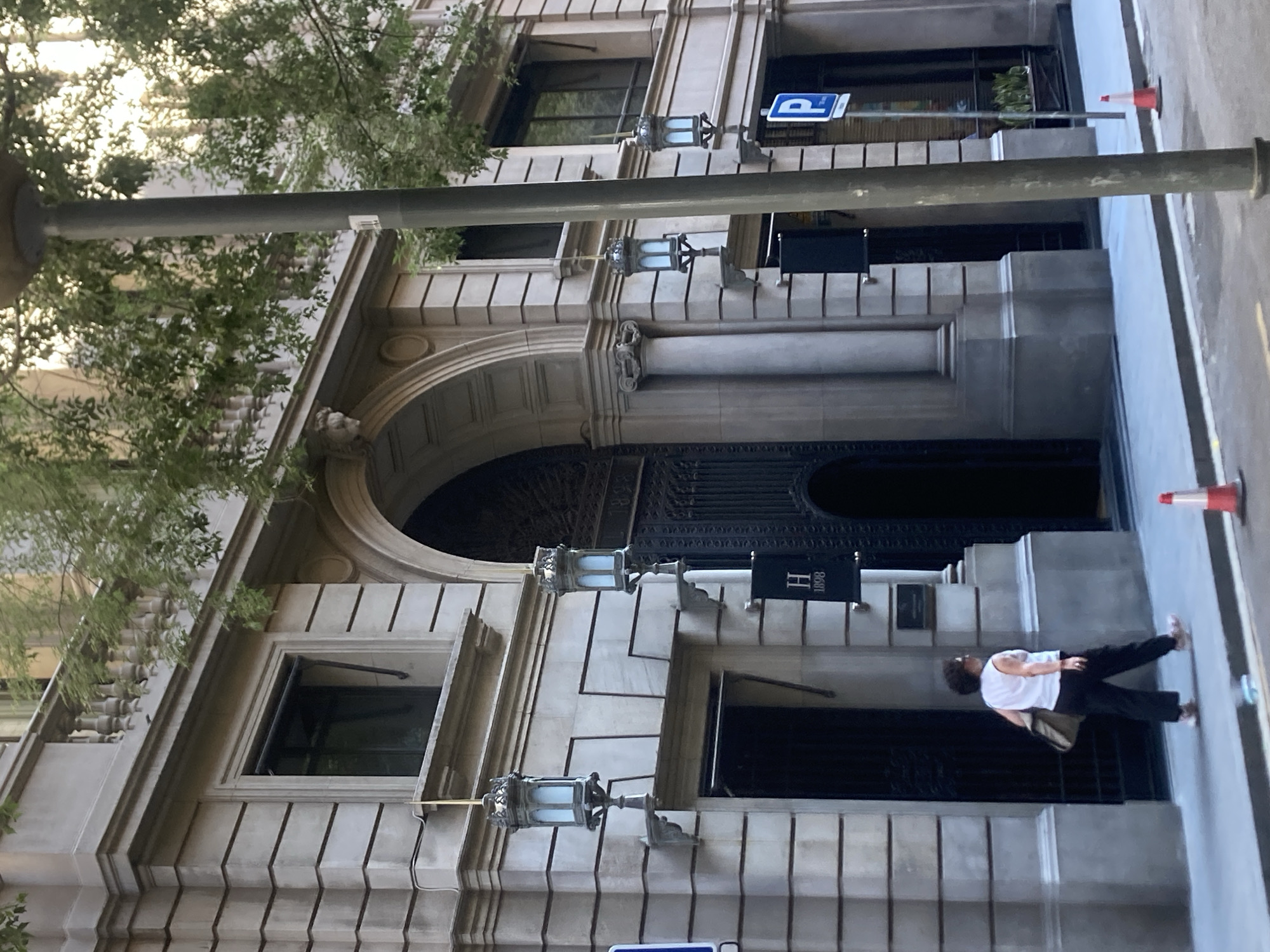 The image shows a person walking on a street in front of a building with classical architecture. The building has a large arched entrance, ornate lamps, and several windows. A sign with the letter "H" and the number "1898" is visible above one of the windows. A blue parking sign is also mounted on the building. A traffic cone is on the sidewalk. The person is wearing a white t-shirt and black pants and appears to be carrying a bag over their shoulder. The scene is lit by natural light, suggesting it is daytime. The architecture and the presence of a parking sign suggest an urban environment, possibly a city center. The time of day appears to be late afternoon or early evening, given the shadows.