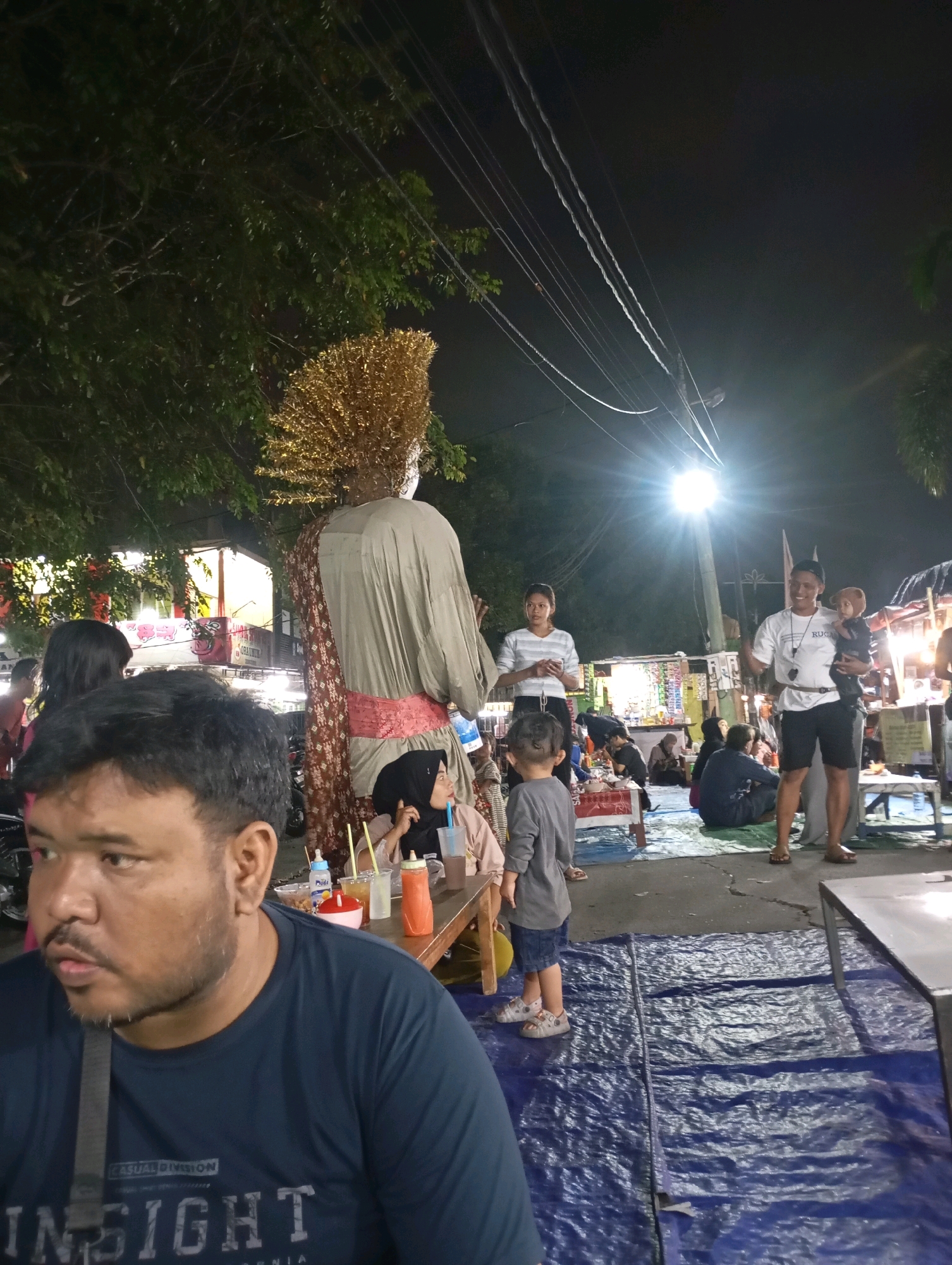 The image depicts a nighttime scene in what appears to be an outdoor market or gathering in Babelan, Indonesia. In the foreground, a man wearing a dark blue t-shirt is seated, looking to his left. To his right, a wooden table holds drinks and what appear to be food items. A large, towering effigy with a golden headdress and a long, tan robe is prominently displayed in the mid-ground, adorned with a red sash. Behind the effigy, a group of people are visible, including a woman in a white striped shirt and black pants, and a man holding a child. The background features various market stalls and a bright street light illuminating the area, casting a warm glow on the scene. Trees with dark foliage frame the upper portion of the image, and power lines are strung across the sky. The ground is covered with blue tarps, suggesting a temporary setup for the event.