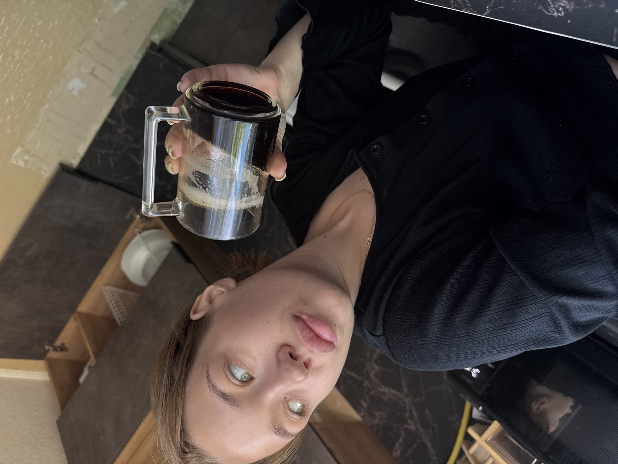 An outdoor, eye-level, medium shot shows a young woman with light brown hair pulled back, looking to her left with light green eyes and full lips. She wears a black, button-up shirt and a thin gold necklace. Her right hand holds a clear glass mug filled with dark coffee, which she brings to her mouth. The background shows a kitchen area with dark countertops and wooden cabinets, some of which are open, revealing dishes and kitchenware. The lighting suggests it is daytime, with natural light entering from the left. The woman is positioned in the foreground, with the kitchen environment serving as a backdrop.