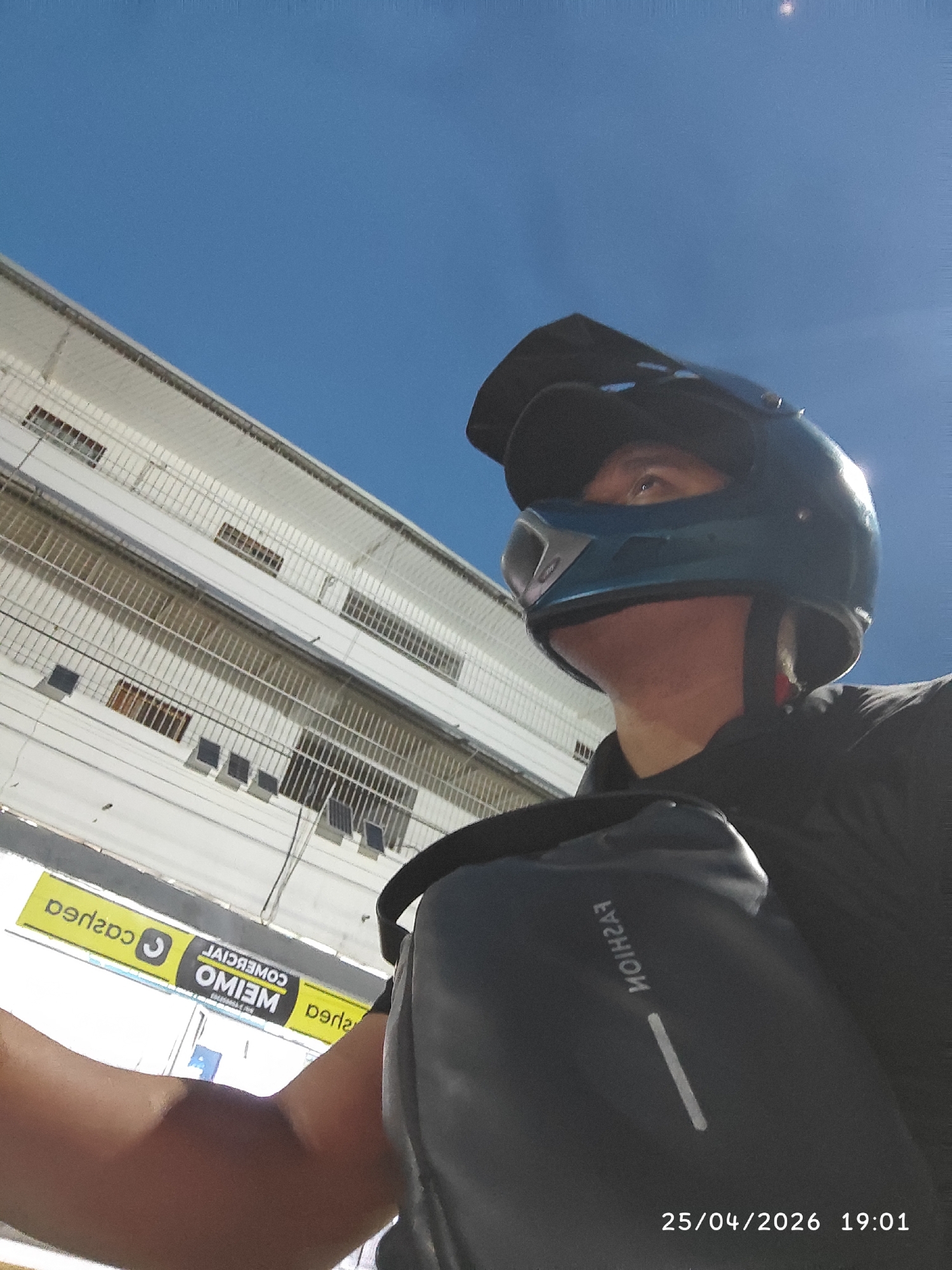 On 25/04/2026 at 19:01, an individual wearing a blue helmet and a black shoulder bag stands in Tinaquillo, Venezuela. The subject is positioned in front of a multi-story white building. A sign is visible with the text "OMIEIM COMERCIAL" and "caspea". The sky is a clear blue.