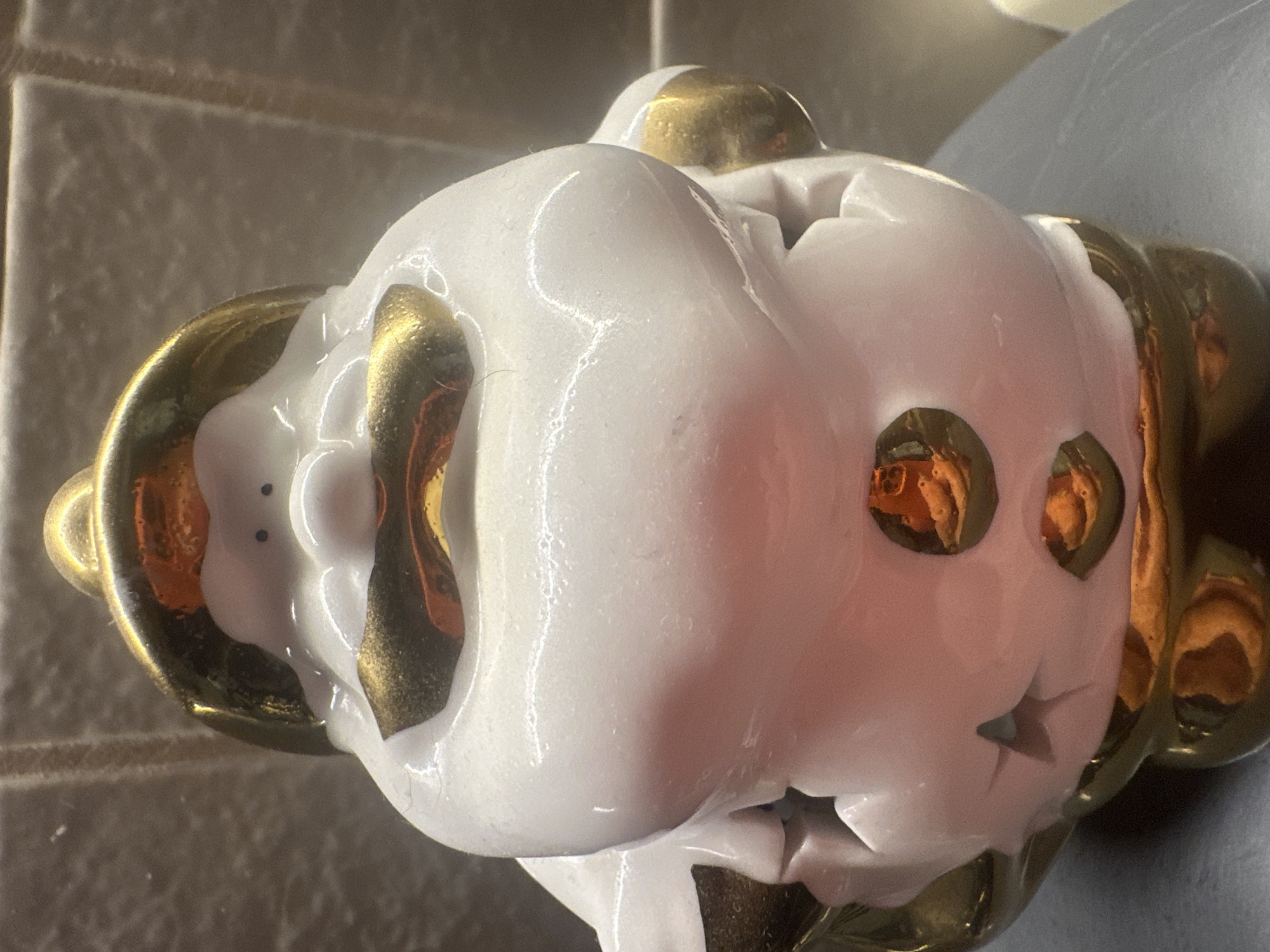 This is a close-up, eye-level shot of a ceramic Santa Claus figurine. The figurine is primarily white with gold accents on its hat, mustache, cheeks, and as decorative buttons on its belly. The background appears to be a tiled wall, with the figurine positioned on a dark surface. There are reflections visible on the gold portions of the figurine, suggesting a light source in front of it. The lighting is somewhat harsh, creating strong highlights and shadows. The overall impression is of a festive, decorative item. The image was taken in the city of Maladzyechna, Belarus.