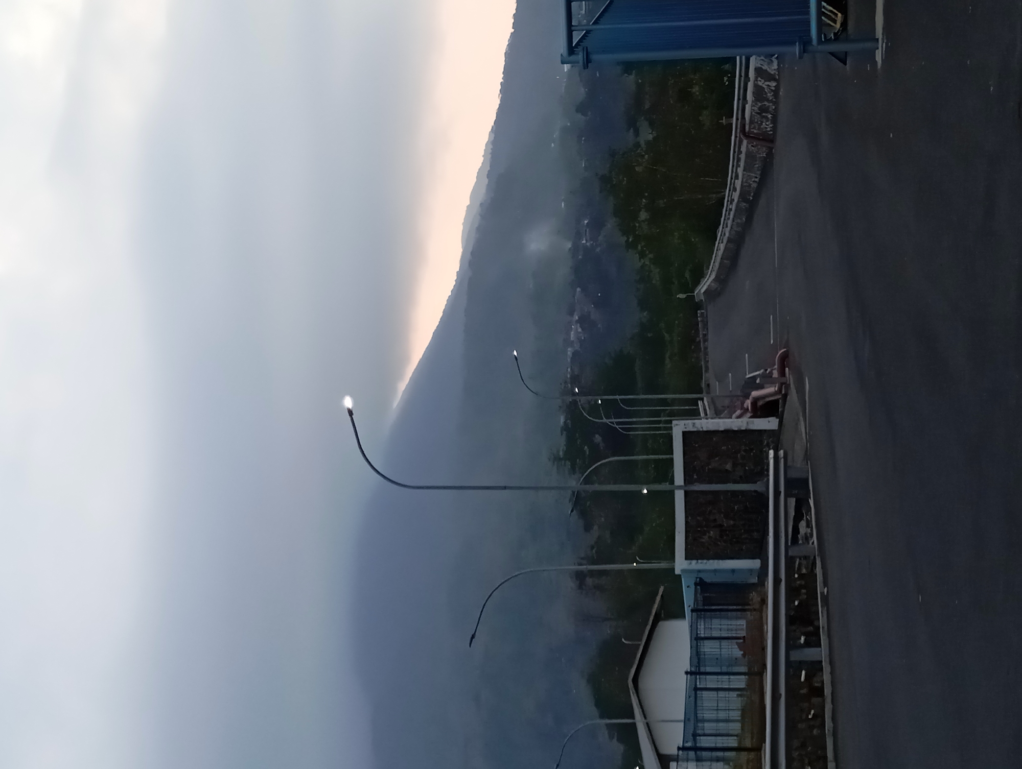 The image depicts an outdoor scene in the city of Sumber Suko, Indonesia, with a prominent mountain in the background partially obscured by low-lying clouds. A paved road curves through the foreground, flanked by streetlights. On the left side of the road, there is a white building with a dark roof, behind a fence. Further down the road on the right, a blue metal structure is visible. A person is seated on the side of the road near a wall. The sky is overcast, suggesting either early morning or late evening.