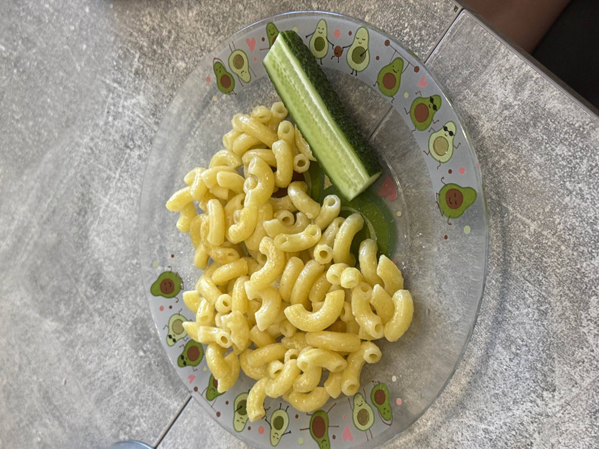 A clear glass plate, adorned with cartoon avocados, holds a portion of macaroni pasta and a slice of cucumber. The plate rests on a gray, textured surface, possibly a countertop or table, in what appears to be a home environment. The lighting suggests it is daytime. No specific location information for Lyubertsy, Russia, is present in the image.