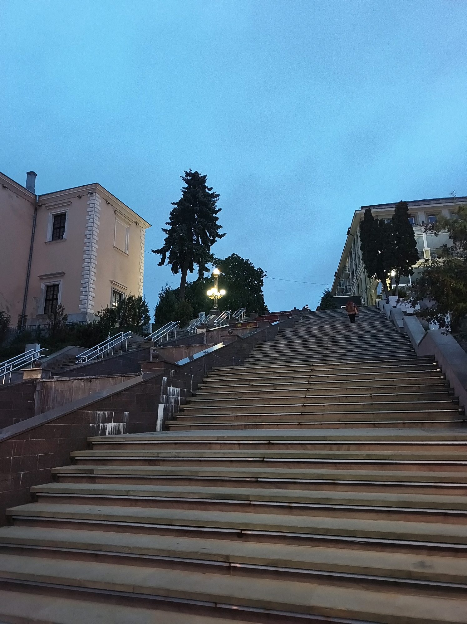 The image captures a wide, stone staircase leading upwards, likely in Ternopil, Ukraine, under a twilight sky. The steps are lit intermittently, creating a sense of depth and leading the eye towards the top. The staircase is flanked by buildings on either side. On the left, a light-colored building with dark windows stands tall, its architectural style suggesting historical significance. On the right, a more modern building with visible windows and some trees in front of it is visible. A lone figure, seen from behind and wearing a backpack, is ascending the stairs in the mid-ground. The overall atmosphere is calm, with the dim lighting and muted colors of the sky contributing to a serene, end-of-day ambiance.