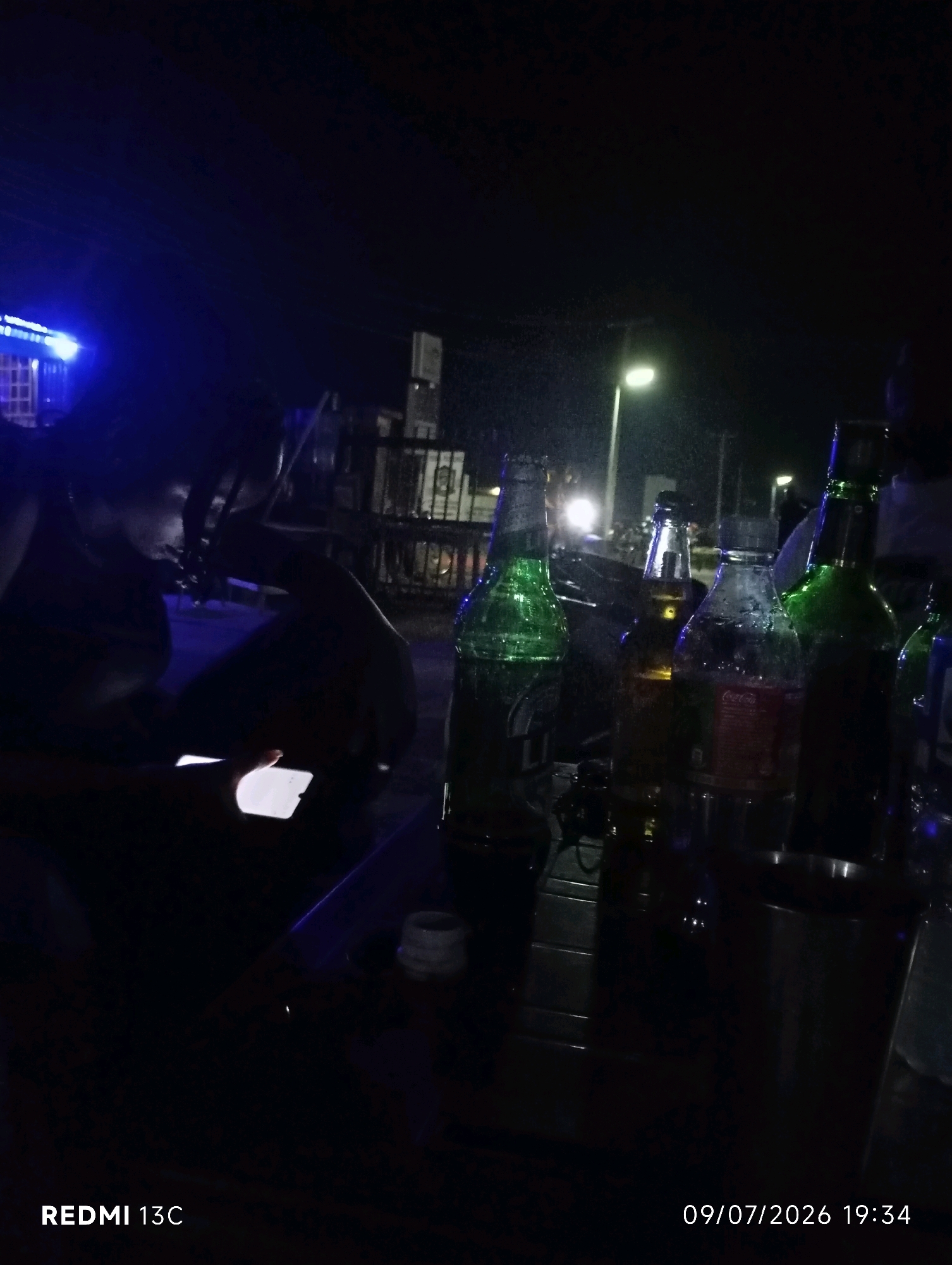 In Shendam, Nigeria, at night, a person is silhouetted in the foreground, holding a glowing smartphone. To their right, several glass bottles, some containing beverages and appearing to be alcoholic, are arranged on a surface. The background is dark, with a street light providing illumination, suggesting an outdoor setting, possibly a roadside stall or a casual gathering place. The image captures a moment of quiet activity, with the soft glow of the phone and the ambient light of the street lamp creating a subdued atmosphere.
