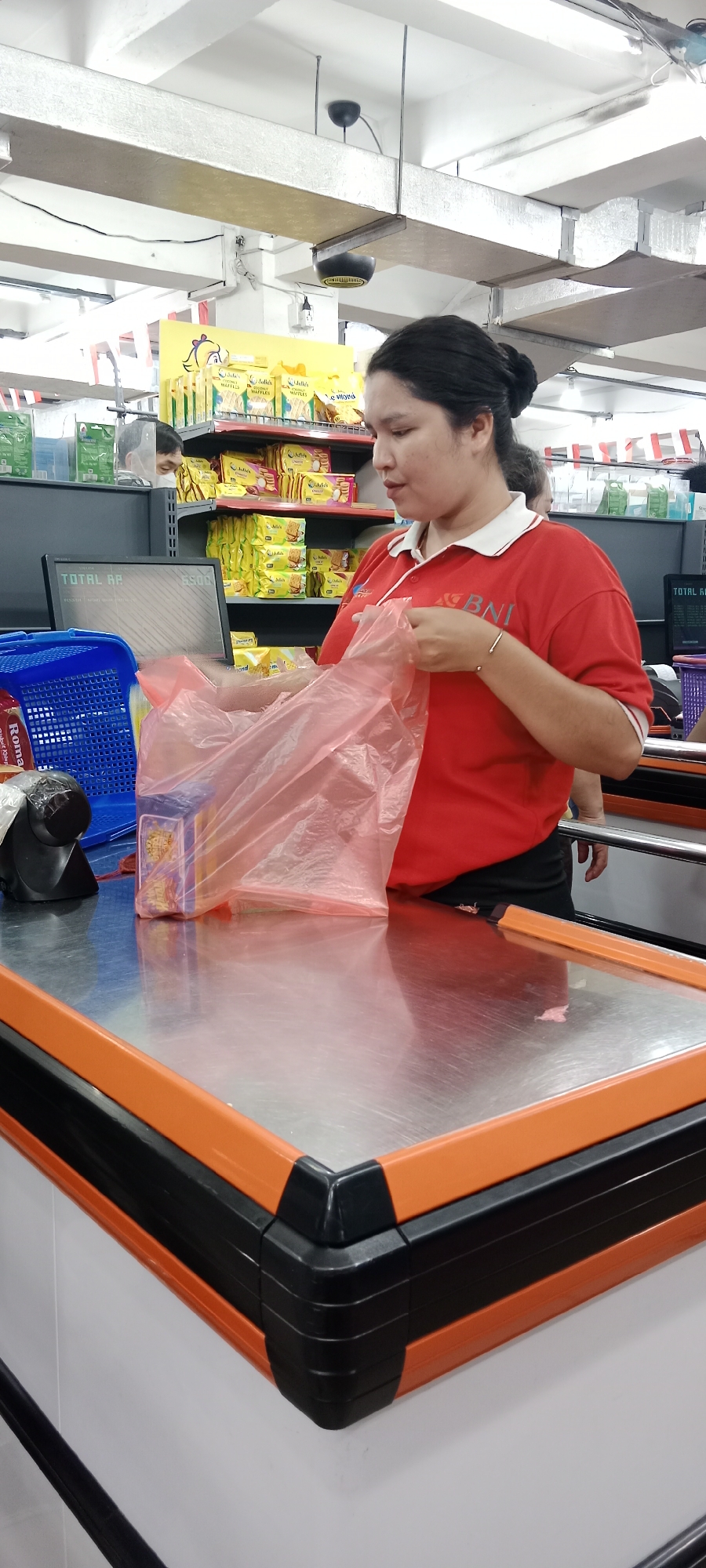 In a supermarket in Tanjung Pinang, Indonesia, a woman wearing a red polo shirt with white collar and accents, a logo on the left chest, and a white stripe around the collar, is standing at a checkout counter. Her dark hair is pulled back in a bun. She is holding a pink plastic bag open with both hands, and appears to be placing items into it. On the counter in front of her, to her left, is a blue plastic basket containing items. A barcode scanner is visible next to the basket. A computer monitor displaying "TOTAL RP" and a numerical value is positioned behind the counter, to the left of the woman. Shelves stocked with yellow and green packaged goods are visible in the background. The checkout counter has a metallic surface with orange and black trim. A second person, partially obscured, is visible behind the counter to the right of the woman. The environment appears to be indoors, with overhead lighting and exposed ceiling infrastructure.