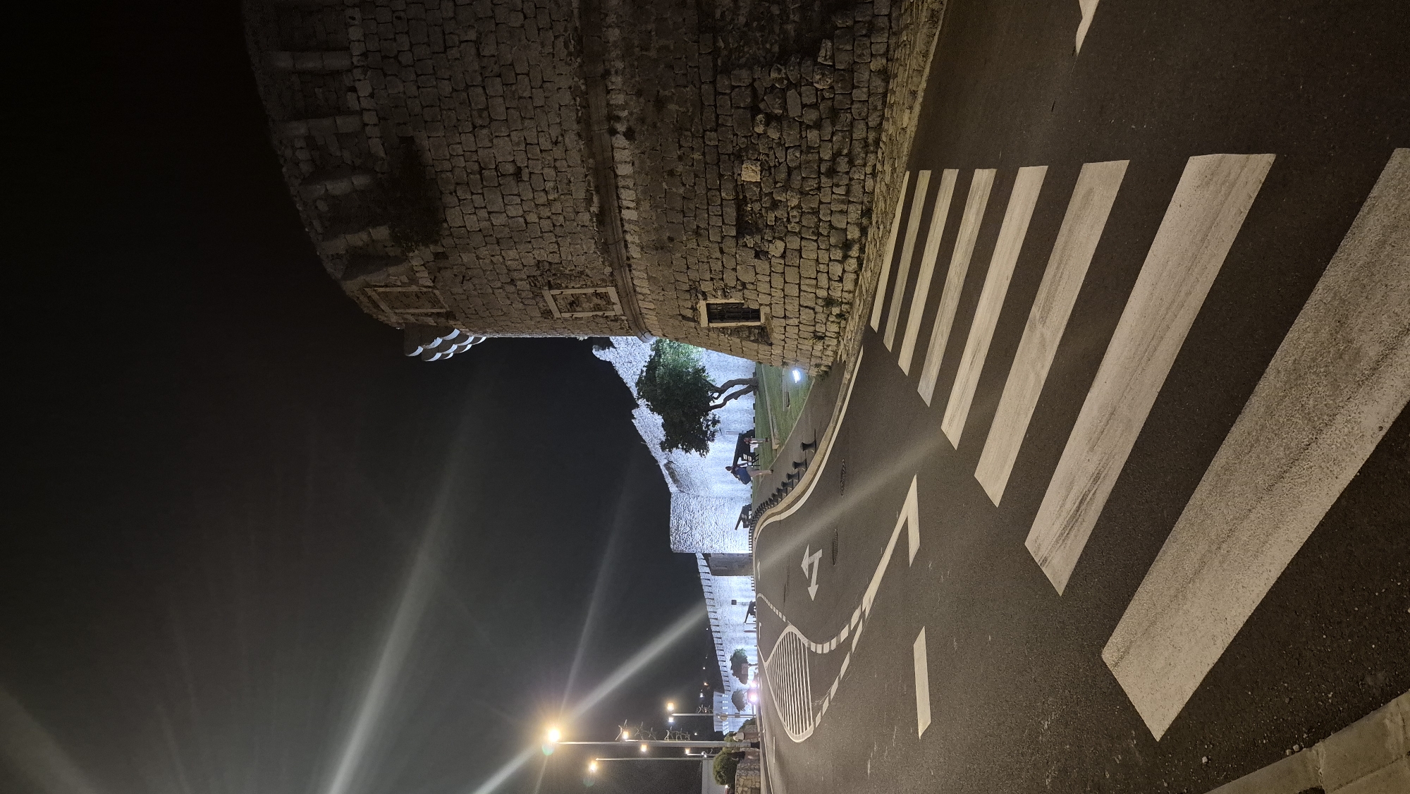 The image shows a nighttime view of an ancient stone wall and a pedestrian crossing. The wall appears to be part of a historical fortification, with rough-hewn stones and arched openings. A tree with dark foliage grows alongside the wall, illuminated by artificial lights. Several streetlights cast beams of light across the scene. The pedestrian crossing is marked with bold white stripes on a dark asphalt road. The road curves gently, with white lane markings and arrows indicating the direction of traffic. In the background, more lights and structures are visible, suggesting an urban or semi-urban environment. The overall impression is one of historical architecture set against a modern urban landscape, with the scene illuminated for nighttime visibility. There is no discernible text in the image. The time of day is clearly night, and the weather appears clear, with no rain or fog evident. The mood is serene, with the stark contrast of light and shadow creating a dramatic atmosphere.