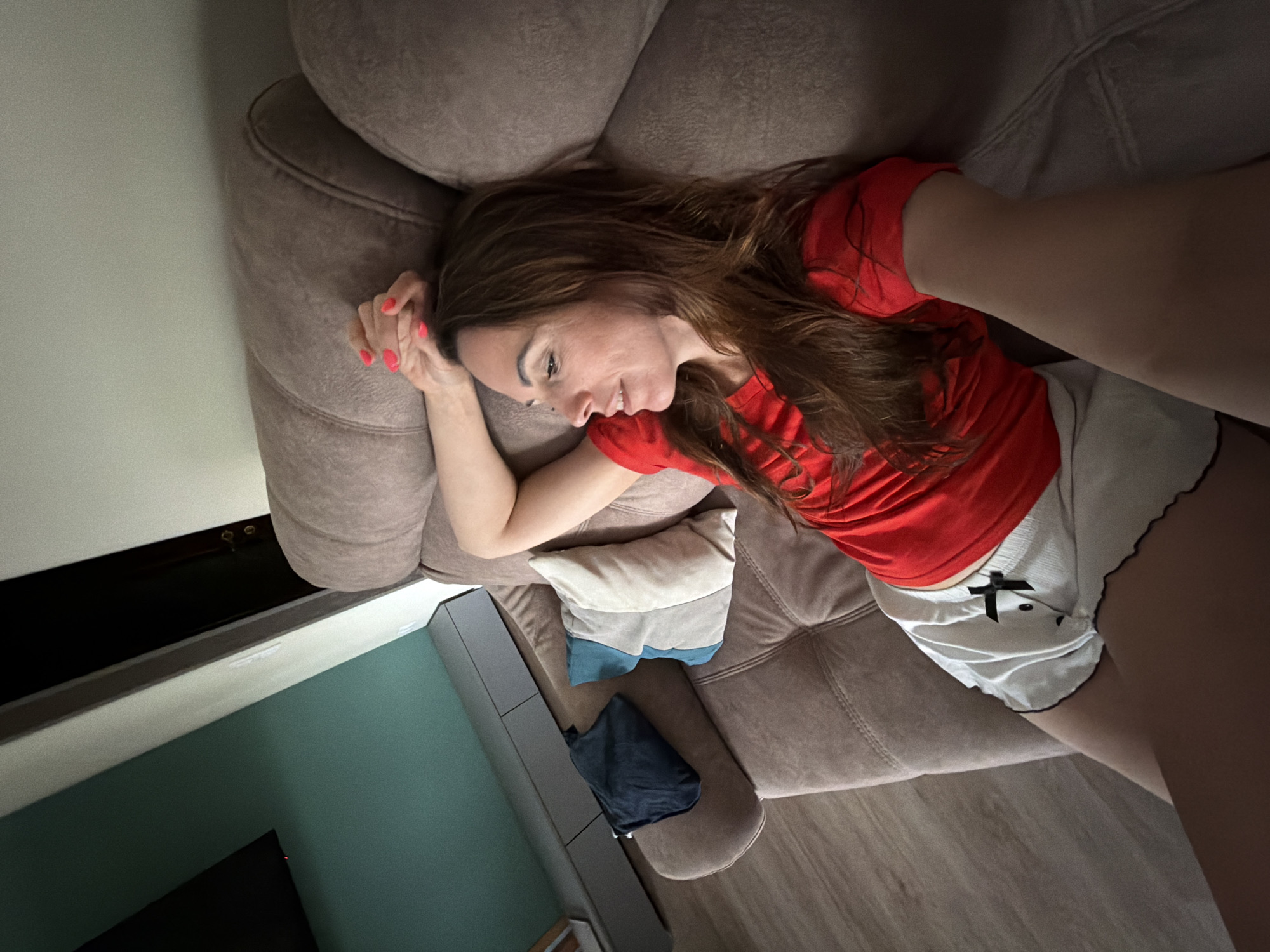 A woman is lying on a brown couch. She is wearing a red shirt and white shorts. Her hair is brown. The couch has two pillows, one of which is gray and blue. There is a black bag on the couch near her. The background shows a wall painted in green and white. The location is likely inside a residence. The location is unlikely to be in Birobidzhan, Russia, as there are no details in the image that point to it.