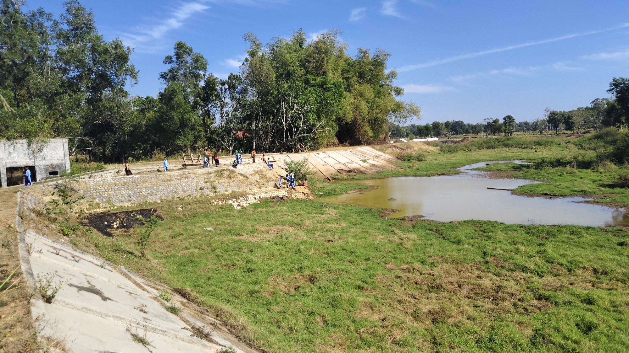 This is a landscape photograph taken outdoors, depicting a natural scene with human presence. The image shows a body of water, possibly a pond or a slow-moving stream, with grassy banks. On the left side of the image, there is a concrete structure, possibly part of a building or retaining wall, with a few people in the vicinity. Further to the right, a group of individuals, including several children, are gathered on a terraced area made of stone and concrete, overlooking the water. The surrounding environment is lush with trees and vegetation. The sky is blue with some scattered clouds. The scene appears to be a public space or a recreational area within the village of Banyubunih, Indonesia, given the context provided. The presence of children suggests it might be a place for leisure or perhaps a school outing. The overall atmosphere is calm and natural, with a hint of human activity.