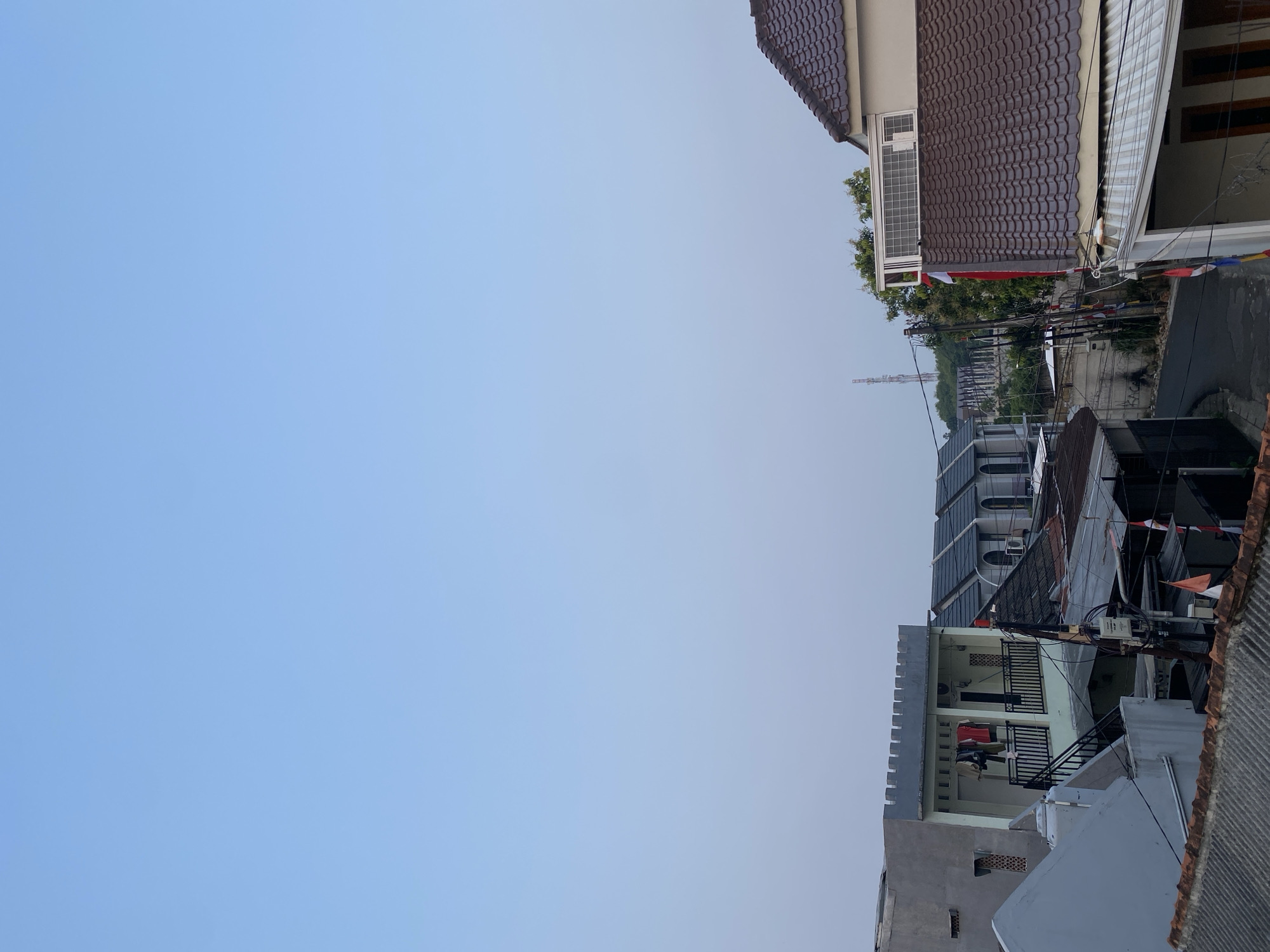 The image depicts a residential area in Tangerang, Indonesia, under a clear sky. In the foreground, several rooftops are visible, with one notably featuring a dark brown, curved tile pattern. To the left, a light gray house with a patterned facade and small windows is partially seen. Adjacent to it is a two-story building with a light green upper floor, featuring a balcony with black railings. Clothes are hanging on a line on this balcony. Behind this building, a row of houses with dark gray, arched roofs is visible, suggesting a planned development. Electrical poles with wires and several small flags are present, some with red and white stripes, possibly indicating a national holiday. In the distance, trees and a transmission tower can be seen. The overall scene suggests a moderately dense urban residential environment.