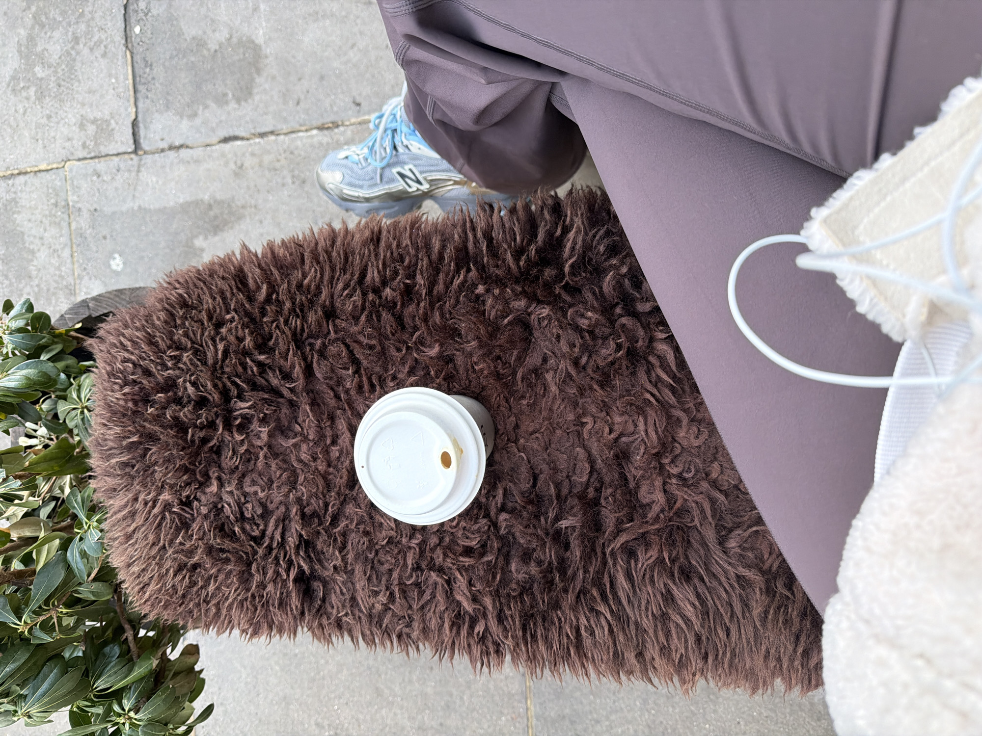 A high-angle close-up captures a street-level scene, featuring a brown furry stool or bench, upon which rests a partially consumed cup of coffee with a white lid. Adjacent to the bench, one can see green foliage, possibly from a shrub or small tree. A person wearing mauve leggings and light blue New Balance running shoes sits or stands nearby. The straps of a white fluffy bag or garment are also visible. The setting appears to be a public space, possibly in London, United Kingdom, as indicated by the urban context with concrete pavement and general environment.