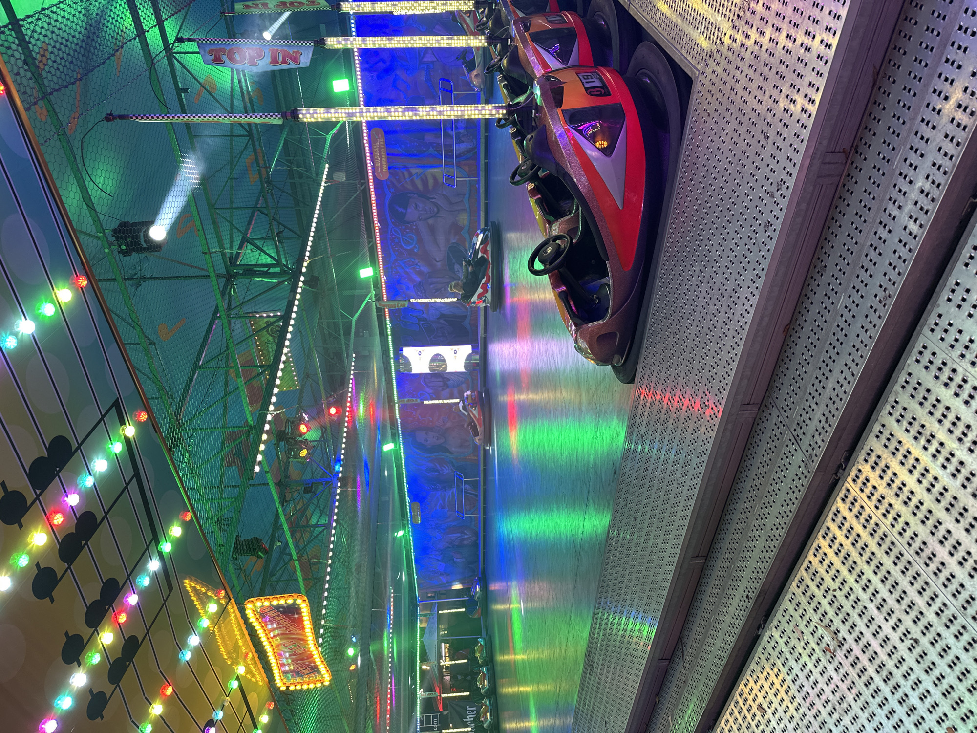 The image depicts an indoor bumper car arena in Bruchhausen-Vilsen, Germany, likely during a fair or amusement event. The arena floor is illuminated with vibrant green and blue lights, reflecting off the polished surface. Several bumper cars are parked in the foreground and mid-ground, with a few occupied by riders. The bumper cars are a mix of red, orange, and black, with sleek, modern designs. In the background, colorful string lights are suspended from the ceiling, and decorative banners with the words "TOP IN" are visible. The walls of the arena are adorned with murals featuring stylized figures. The overall atmosphere is festive and energetic, suggestive of a fun amusement park attraction.