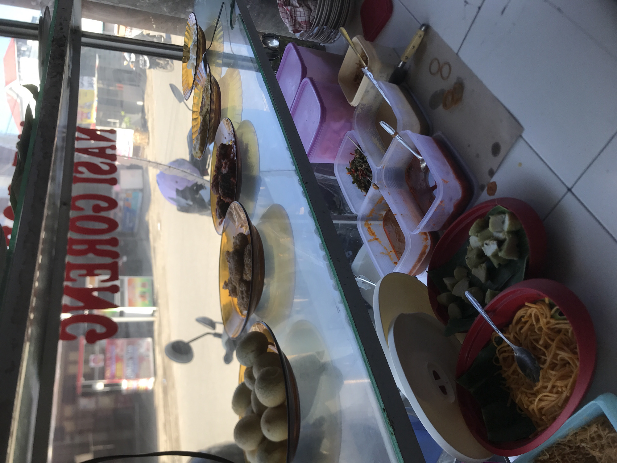 The image shows a display case filled with various food items, likely in Lhokseumawe, Indonesia, given the reflection of a sign that reads "NASI GORENG" (Fried Rice), a popular Indonesian dish. The display case contains plates with fried snacks, meat dishes, and round white balls, along with plastic containers filled with various condiments and side dishes like tofu, spices, and noodles. The arrangement suggests this is a street food vendor preparing or displaying items for sale. Utensils like spoons and knives are also visible near the food containers. The surrounding environment reflected in the glass shows a street scene with motorcycles and buildings. The image is taken at an angle, and some of the items are partially cut off by the frame.