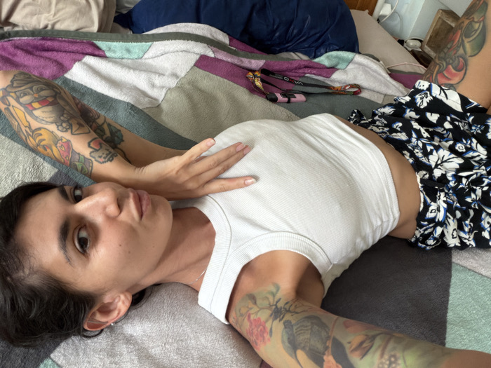 A single individual is depicted lying supine on a bed in what is identified as Nusa Dua, Indonesia. The subject is a female, appearing to be a young adult, with dark hair and a direct gaze towards the camera. She is wearing a white ribbed crop top and black shorts with a white and blue floral pattern. Both of her arms are extensively covered with colorful tattoos featuring various designs, including a figure with a hat on her right forearm and a bird with floral elements on her left bicep. Her right hand is placed on her chest. The bed is covered by a multi-colored blanket or bedspread in a patchwork or striped pattern, incorporating shades of white, gray, purple, and teal. A dark blue pillow is visible behind her head. In the upper right background, a strap-like object, possibly a leash or bag strap, rests on the blanket. The scene is illuminated by ambient indoor lighting.