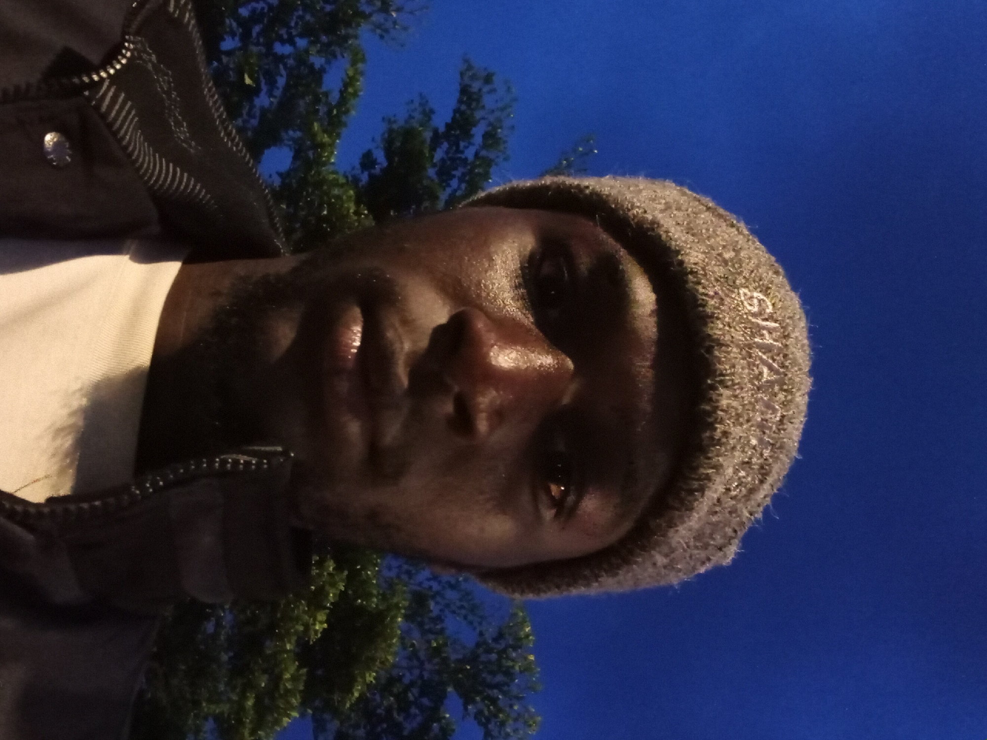 A man wearing a gray beanie and a black jacket poses for a selfie at night. He has a light-colored shirt underneath his jacket. The background is a dark blue sky with trees visible. The beanie has the word "GHANA" embroidered on it. The scene appears to be outdoors in a city, possibly Bukuru, Nigeria, given the context.