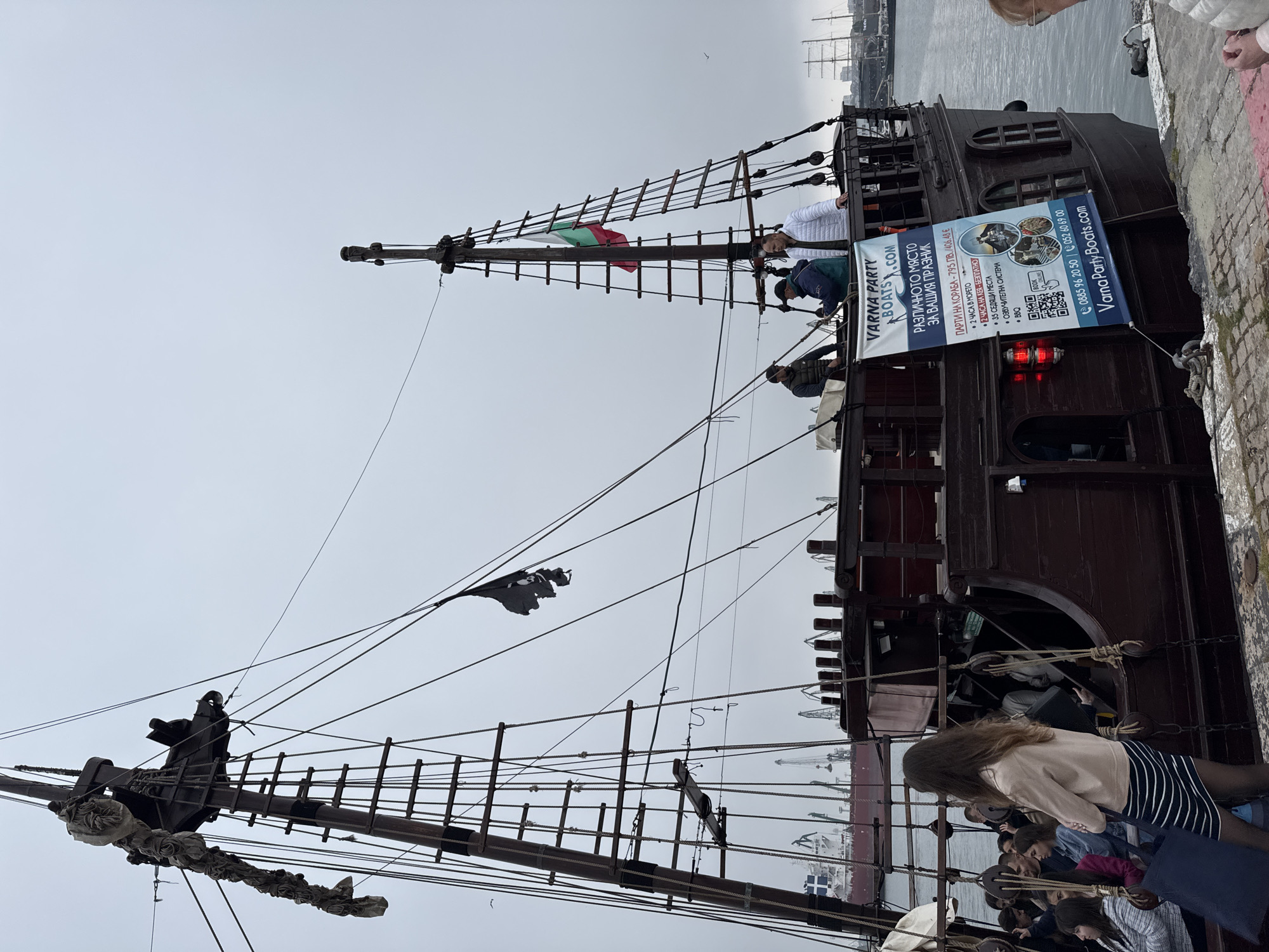The image captures a scene at the Varna harbor in Bulgaria, dominated by a wooden ship adorned with a pirate flag and the Bulgarian flag. People are visible on the deck of the ship and near the edge, with one woman near the edge with a skirt and jacket, and more people can be seen sitting in the background. The ship has masts with ladders and rigging. A large banner on the ship advertises "Varna Party Boats.com" and provides details about parties on the ship, including prices, amenities, and contact information. Cranes and other ships are visible in the background on the water, suggesting the presence of port activity. The sky is overcast, indicating a gray, cloudy day.
