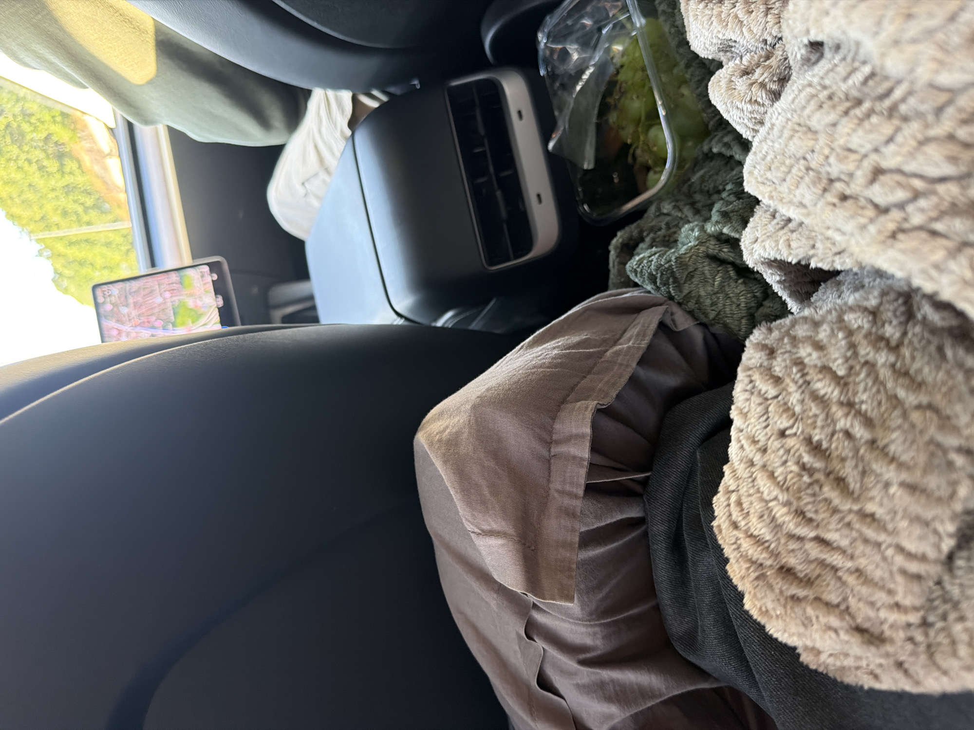 The image shows the interior of a car, likely a Tesla, from a rear-seat perspective. In the foreground, several soft blankets are piled up, with a brown, textured blanket in the very front. Behind the blankets, a container of grapes is visible. Further back, the car's center console with air vents is prominent, and to the left, a portion of the car's entertainment screen is visible, displaying a map with pink and green lines. A person is partially visible in the background, wearing a green hoodie and light-colored pants, looking out the window. The exterior scenery suggests a sunny day with greenery visible through the car windows. The specific location in Gothenburg, Sweden cannot be determined from the visual information provided in the image.