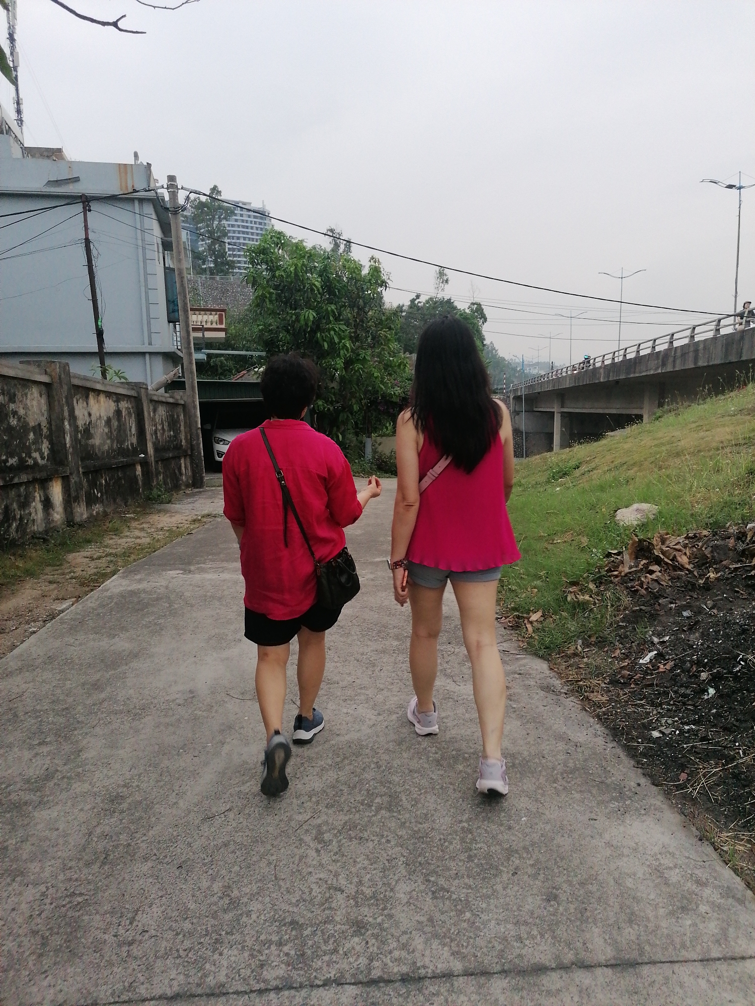 Two women are walking away from the viewer on a concrete pathway. The woman on the left is wearing a red shirt, black shorts, and dark sneakers, with a black purse slung across her shoulder. The woman on the right has long dark hair and is wearing a pink sleeveless top, grey shorts, and light pink sneakers. To the left of the pathway is a concrete wall, and on the right is a grassy area with some dead leaves. A bridge spans the background, and buildings and trees are visible in the distance. The image appears to be taken in an urban or suburban setting within Bai Chay Ward, Vietnam.