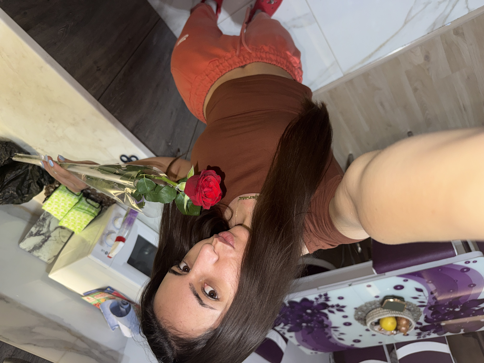 A selfie shot from above shows a woman holding a red rose in a clear plastic wrapper. She has long dark hair and wears a brown t-shirt and orange sweatpants. Her feet, clad in red slippers, are visible at the top of the frame. The background includes a kitchen counter with a white microwave, cleaning supplies, and a coffee table with a white patterned surface and purple legs. The location appears to be inside an apartment. There is no evidence to confirm the image was taken in Plovdiv, Bulgaria.
